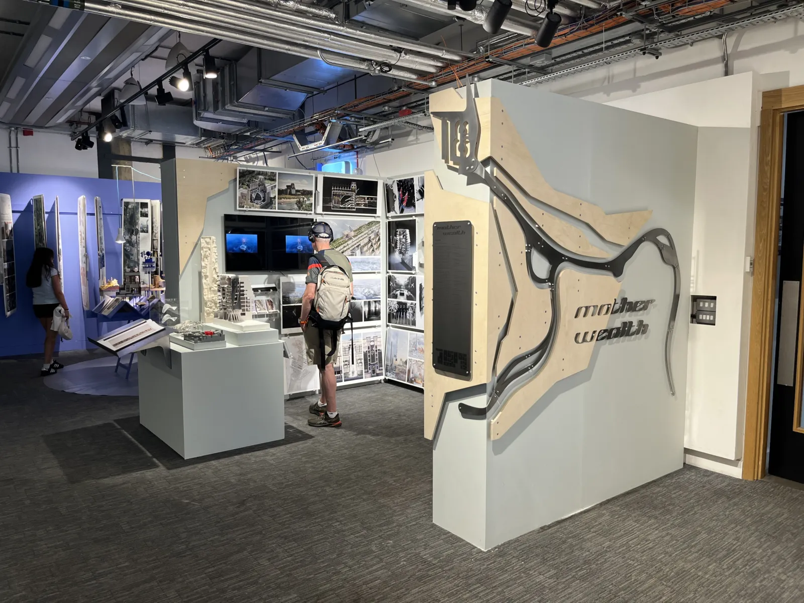 Architecture exhibition displaying concept models, drawings, photographs and digital media related to a large infrastructure project. Visitors view exhibits arranged around freestanding display panels, while a sculptural wall-mounted installation featuring the words “mother wealth” occupies the foreground.