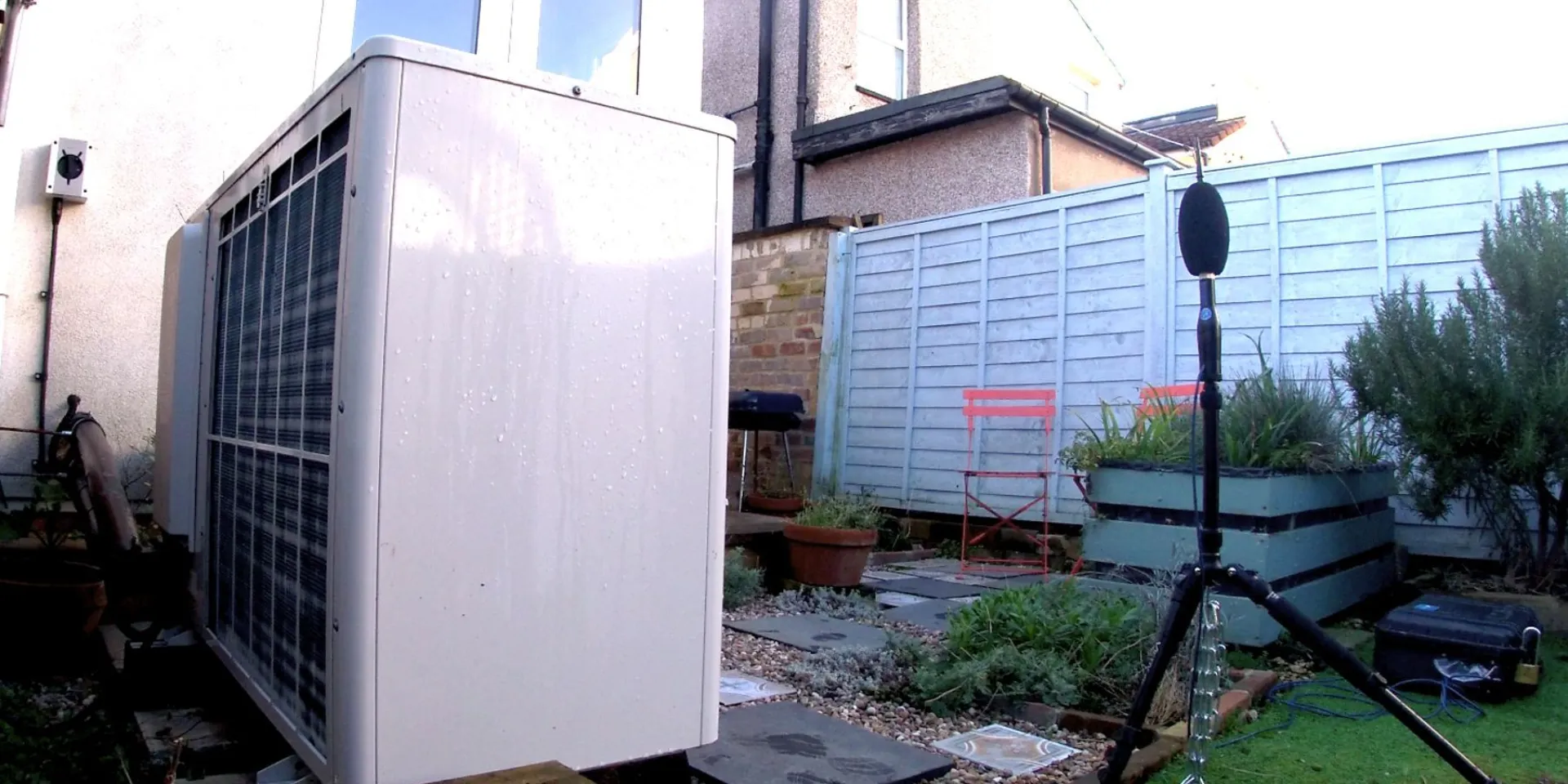 Images show a large, white rectangular box, which is an air source heat pump, in the garden next to a house. There is recording equipment positioned to record the sound coming from the air source heat pump