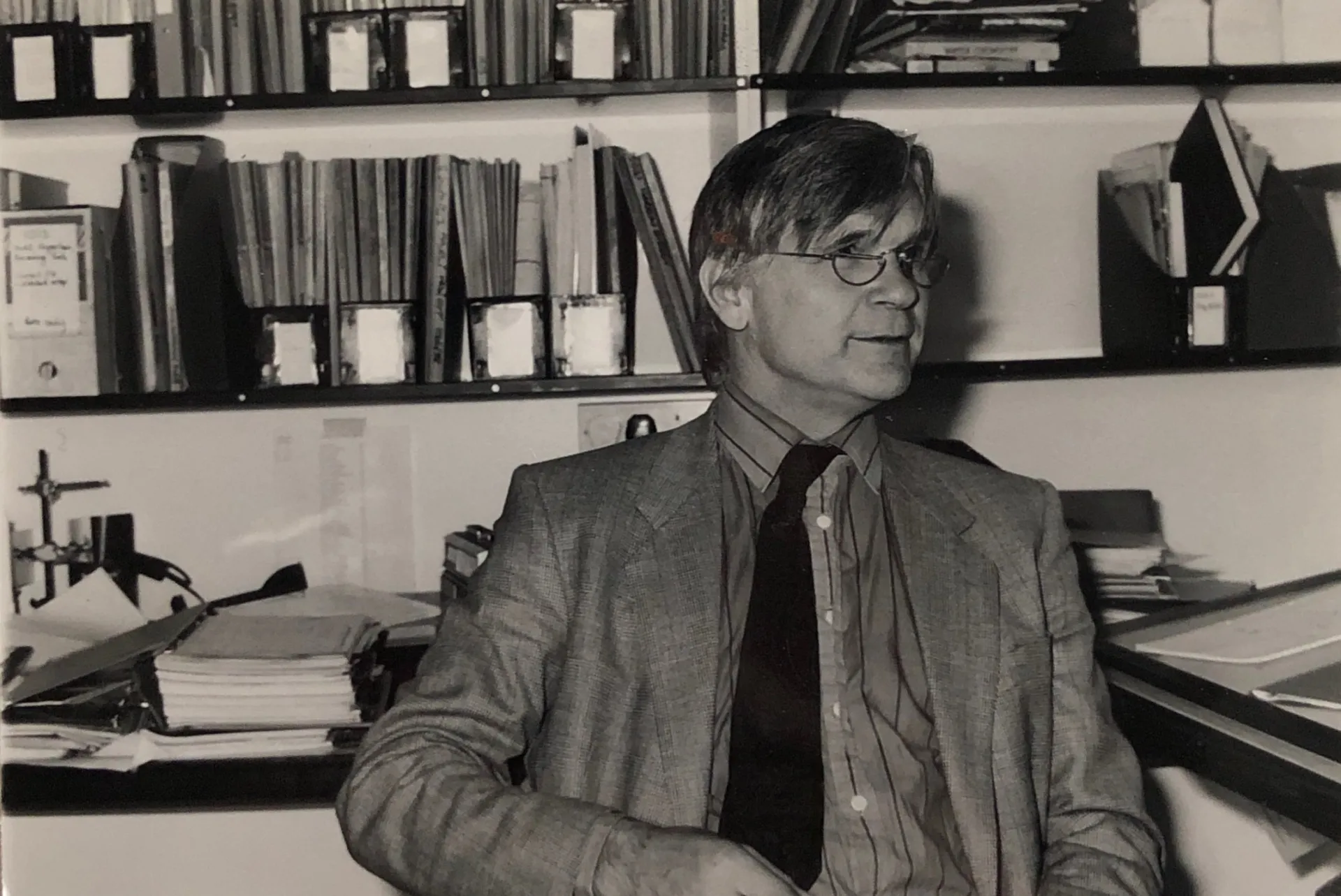 Black and white photo of Max in a jacket and tie, sat in the office in front of a book shelf.