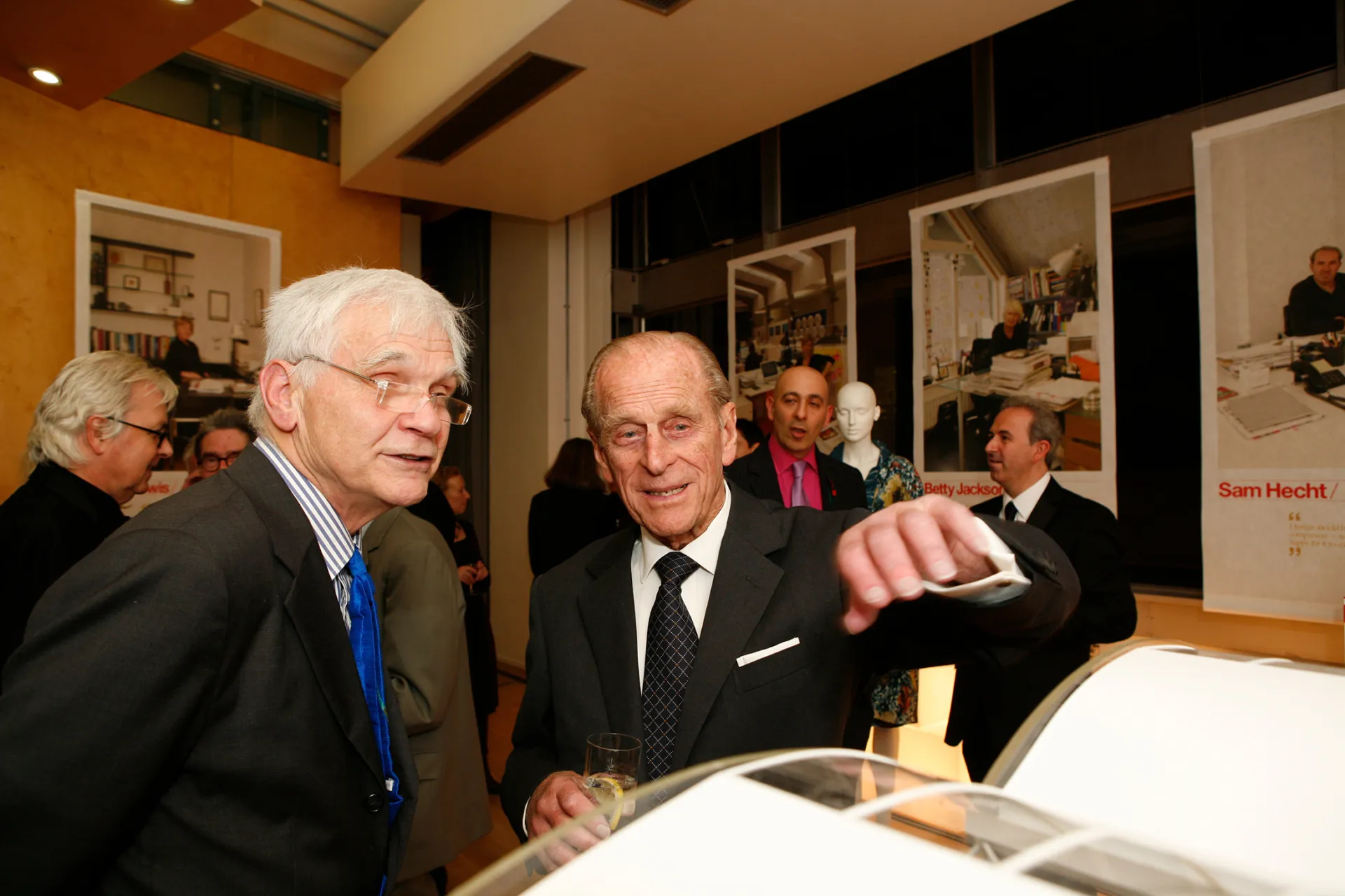 Max talking to Prince Philip at an event, both wearing suits and discussing an Architect's model in front of them.