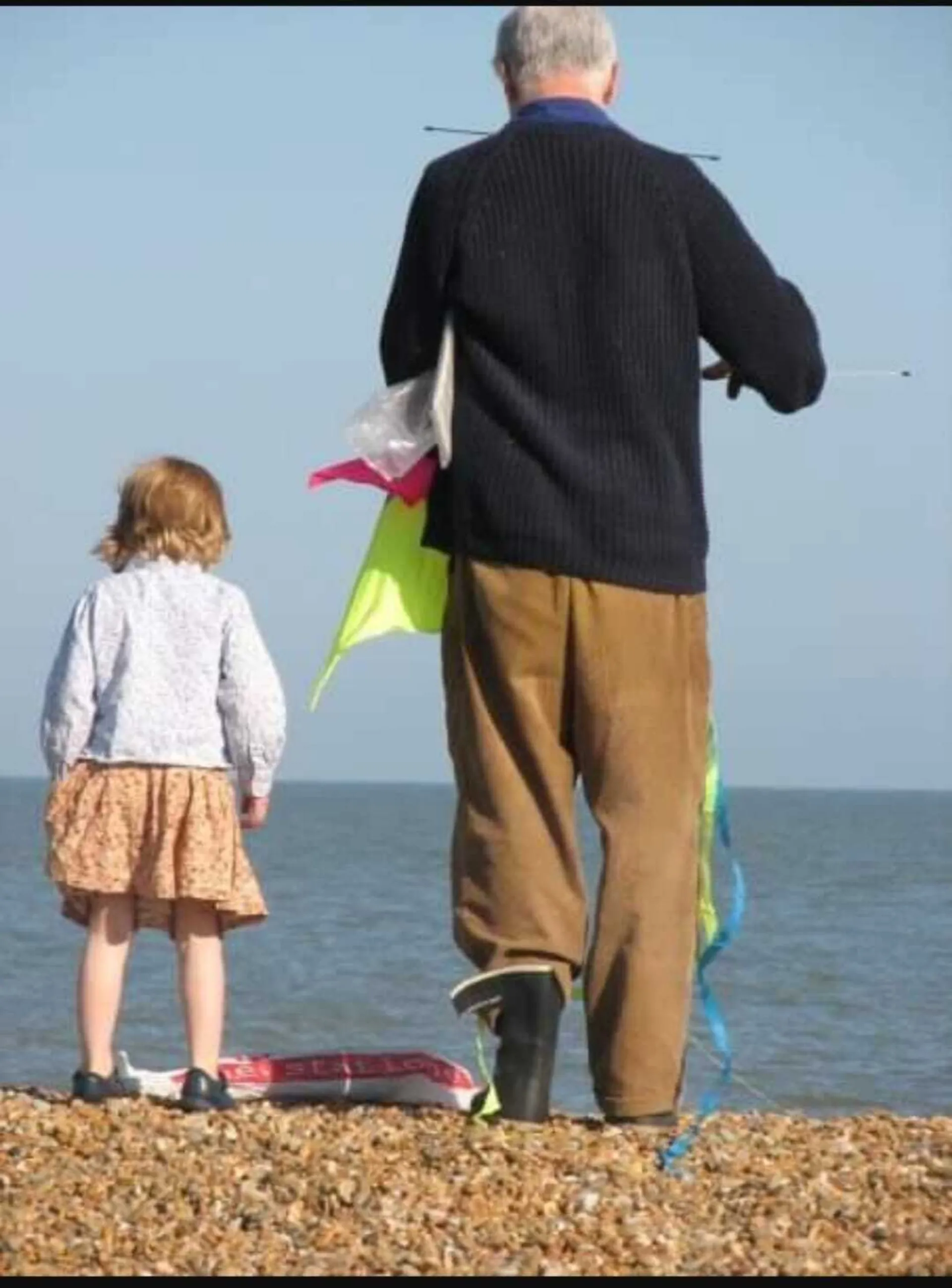 Colour photograph of Max as an elderly man in a dark jumper, trousers and wellington boots. standing next to a young child in a dress and white jumper. Both are facing away from the camera looking towards the sea. Max is holding a kite.