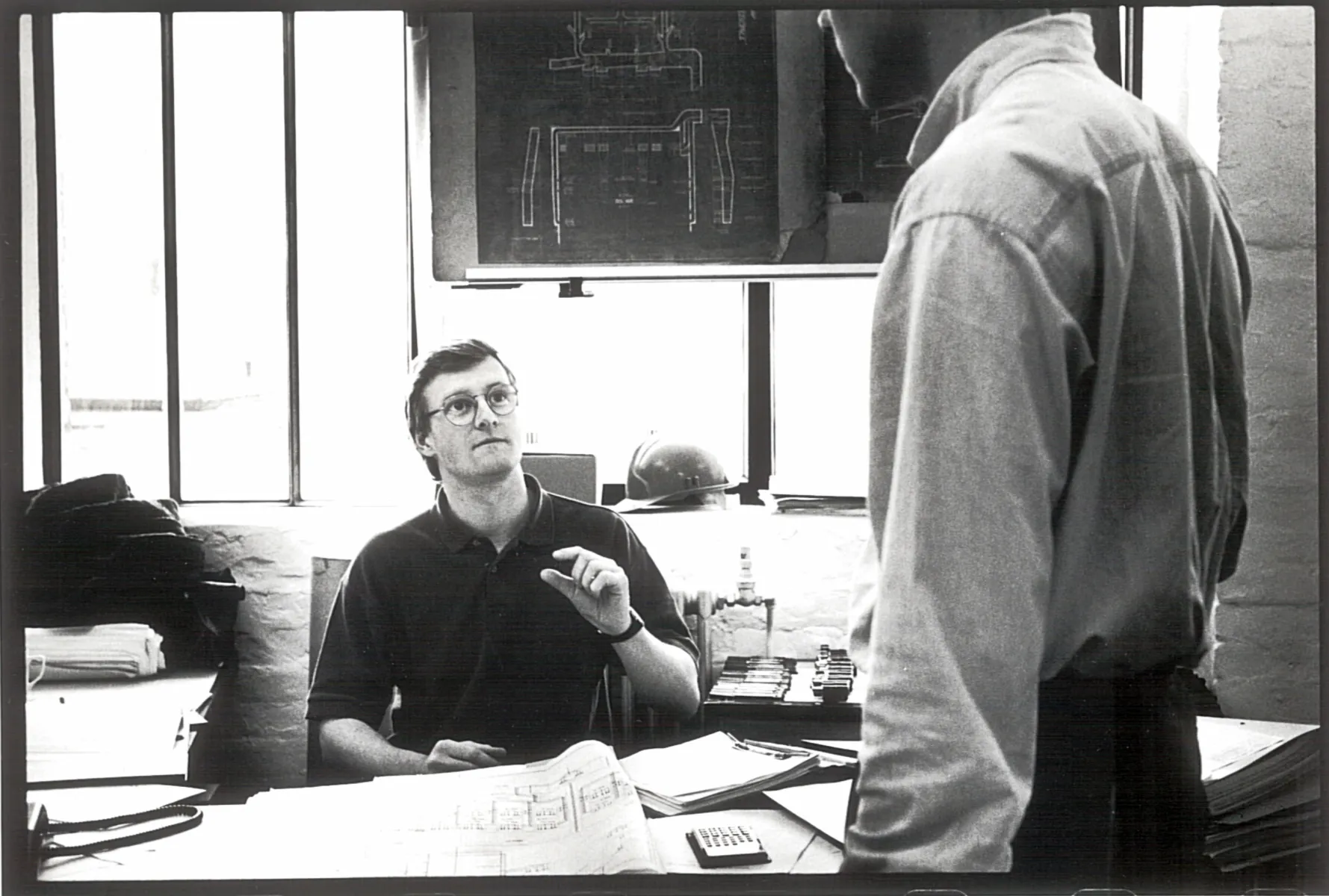 black and white showing man seated and speaking to a standing man with a lot of paper on this desk