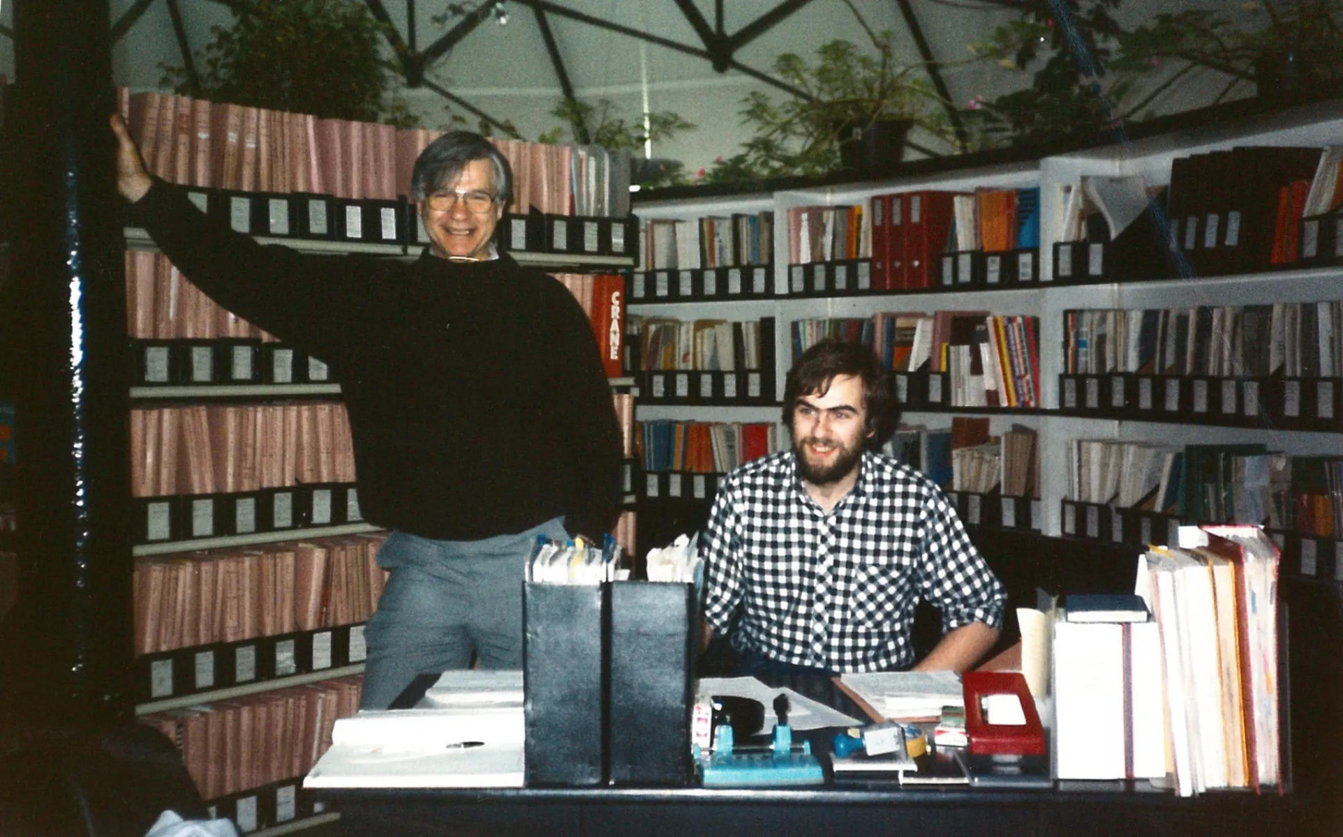 Two men one standing and one seated with files behin them.