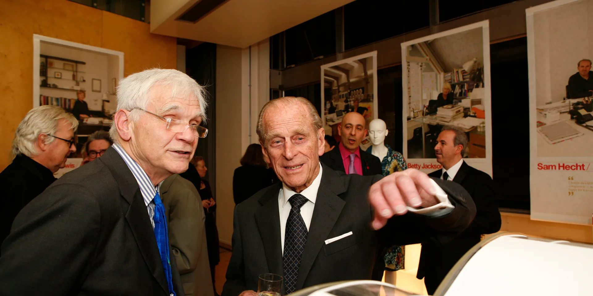 Max talking to Prince Philip at an event, both wearing suits and discussing an Architect's model in front of them.