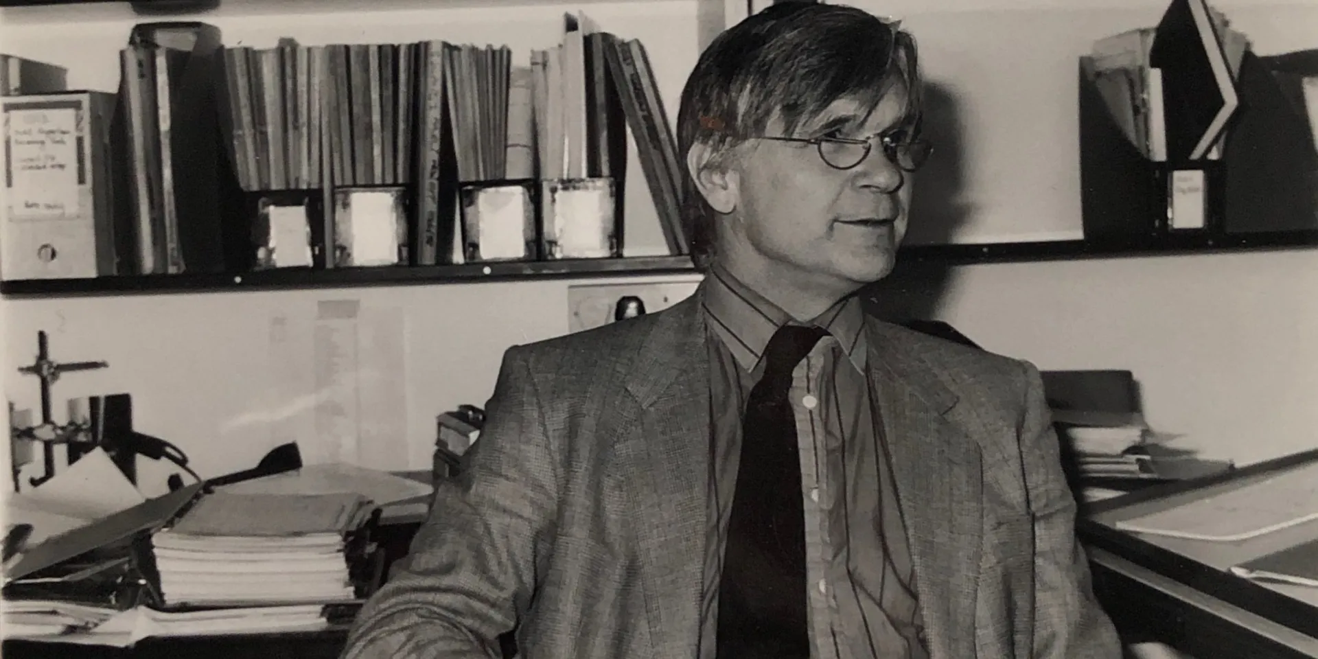 Black and white photo of Max in a jacket and tie, sat in the office in front of a book shelf.