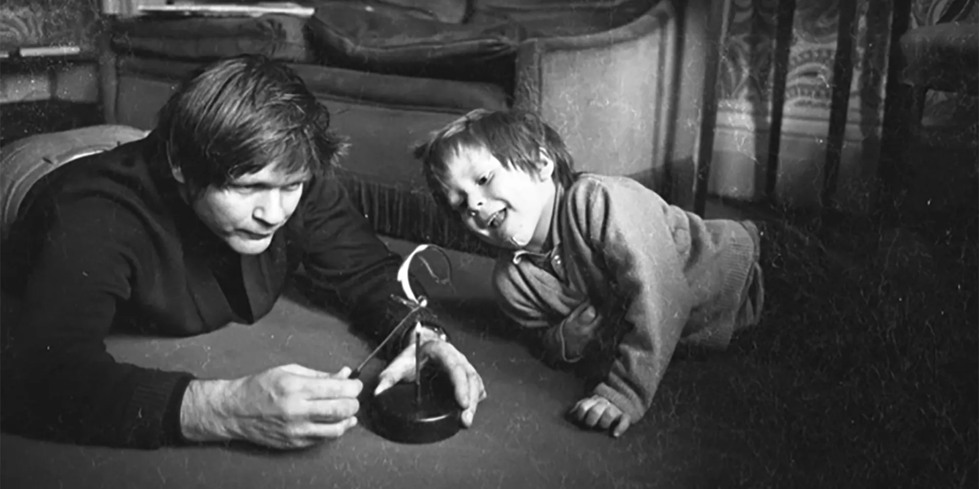 1970s black and white photo of Max and his young son Finn, both on the living room carpet. Max looks serious and is demonstrating the operation of some gadget to Finn, who is laughing.