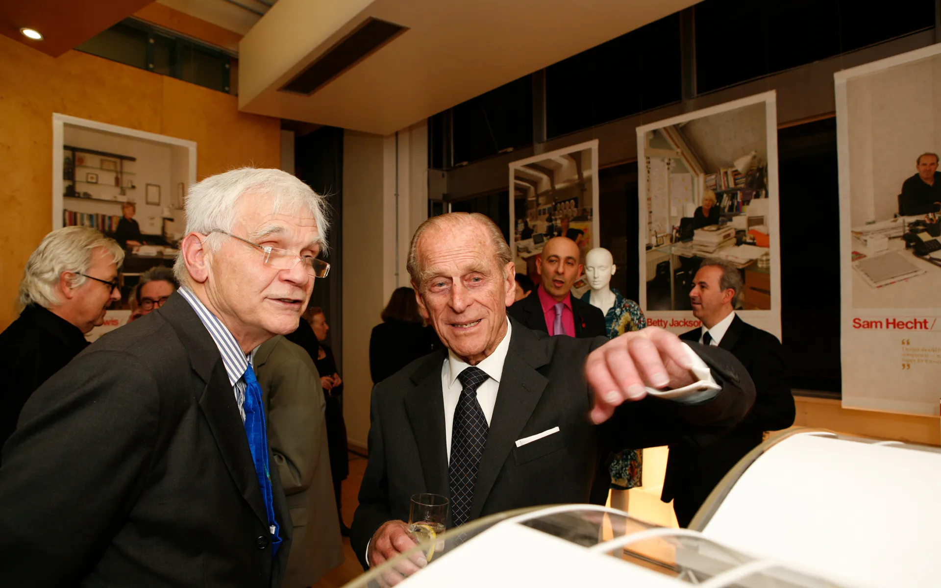 Max talking to Prince Philip at an event, both wearing suits and discussing an Architect's model in front of them.