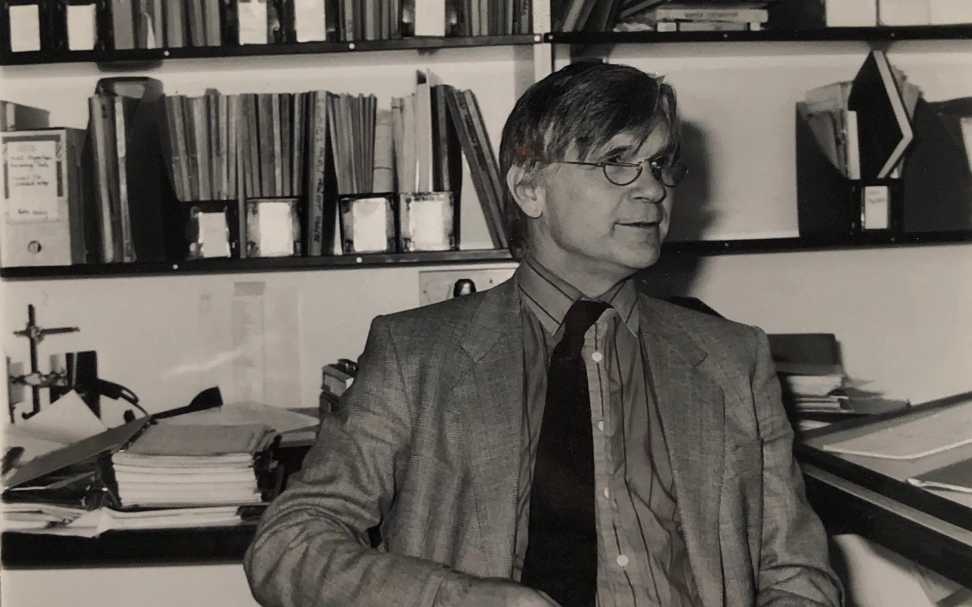 Black and white photo of Max in a jacket and tie, sat in the office in front of a book shelf.