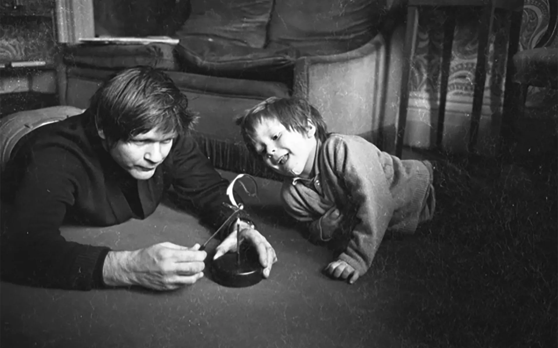 1970s black and white photo of Max and his young son Finn, both on the living room carpet. Max looks serious and is demonstrating the operation of some gadget to Finn, who is laughing.