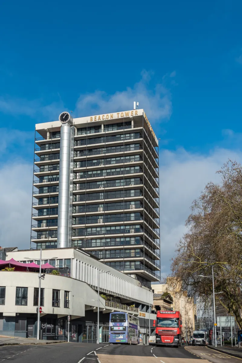 A tall modern building named "Beacon Tower" with multiple balconies and large windows standing under a clear blue sky. There is a clock on a cylindrical structure attached to the side of the building. Trees are visible on the right side of the image.