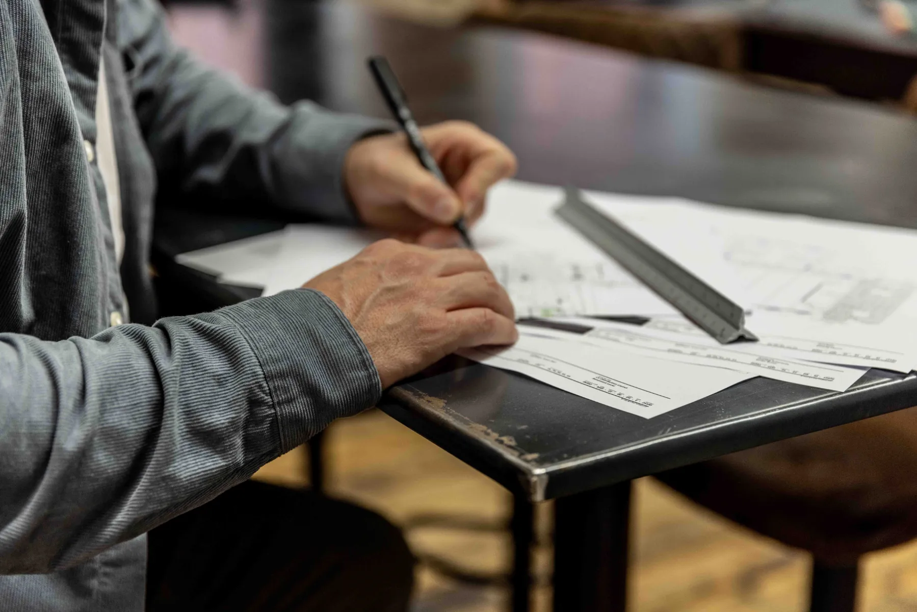 Close up showing hands of someone drawing at a desk with pen, paper and ruler.