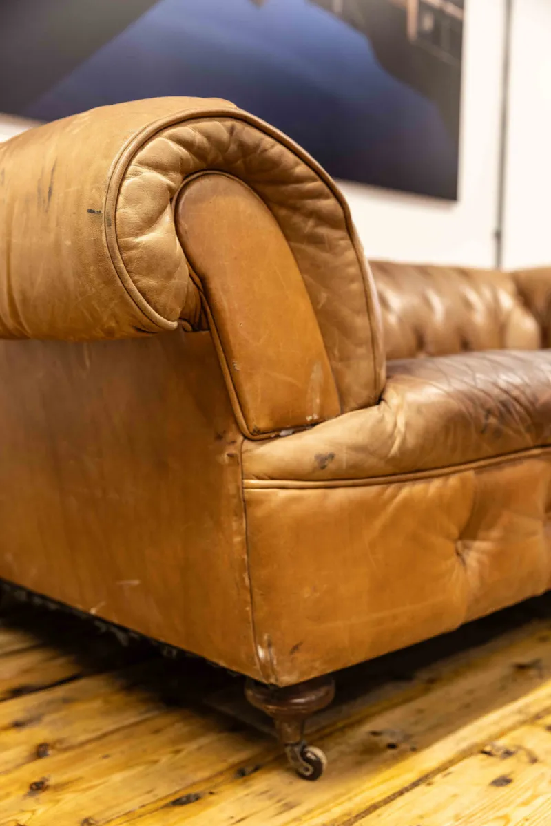 Close up showing the arm of a worn, tan leather sofa on a wooden floor.