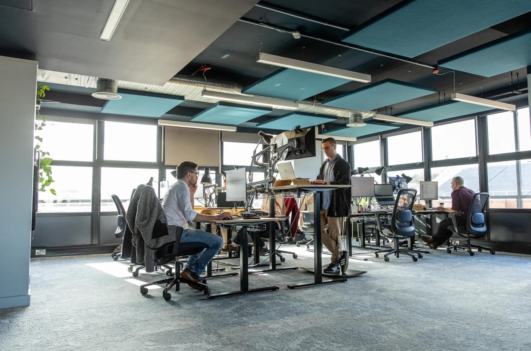 Internal view of the office, with people sitting and standing at desks, and windows in the background