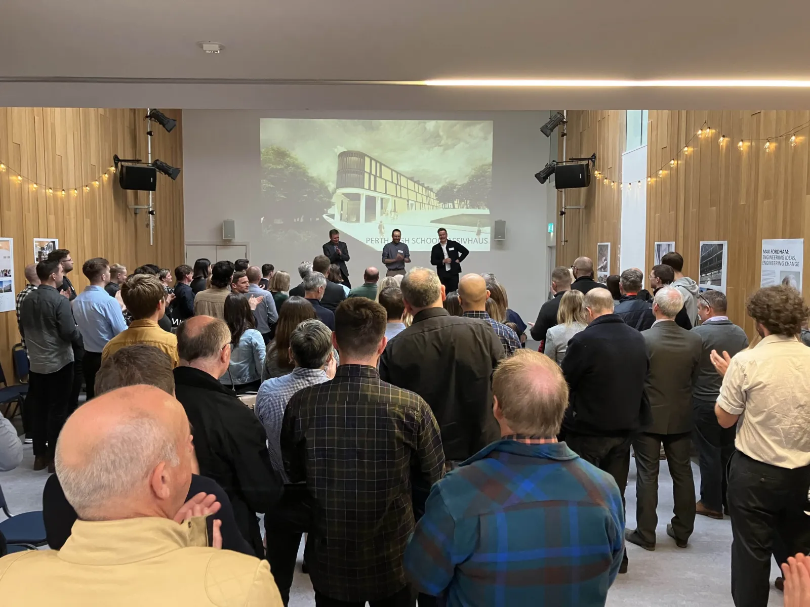 Lots of people in an event space with three people on stage making a speech