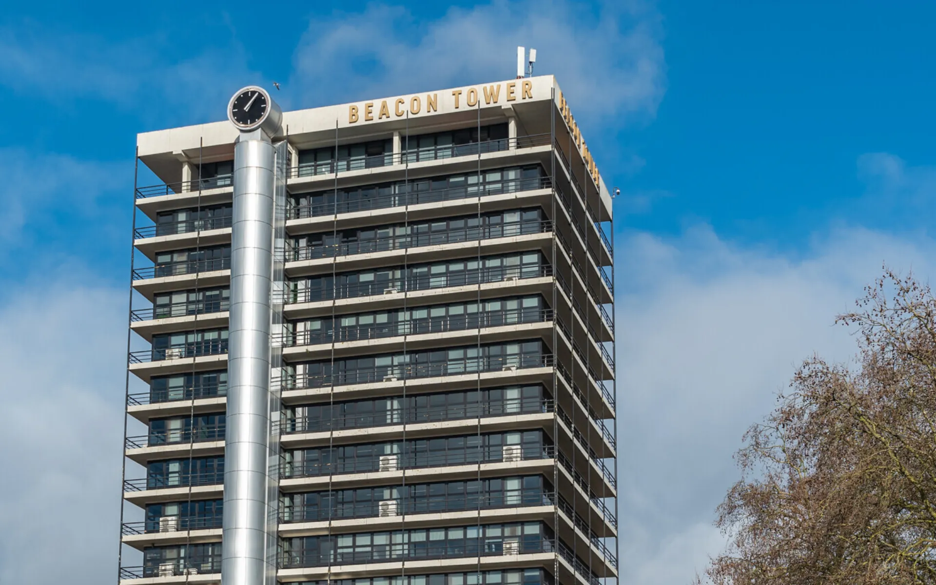 A tall modern building named "Beacon Tower" with multiple balconies and large windows standing under a clear blue sky. There is a clock on a cylindrical structure attached to the side of the building. Trees are visible on the right side of the image.