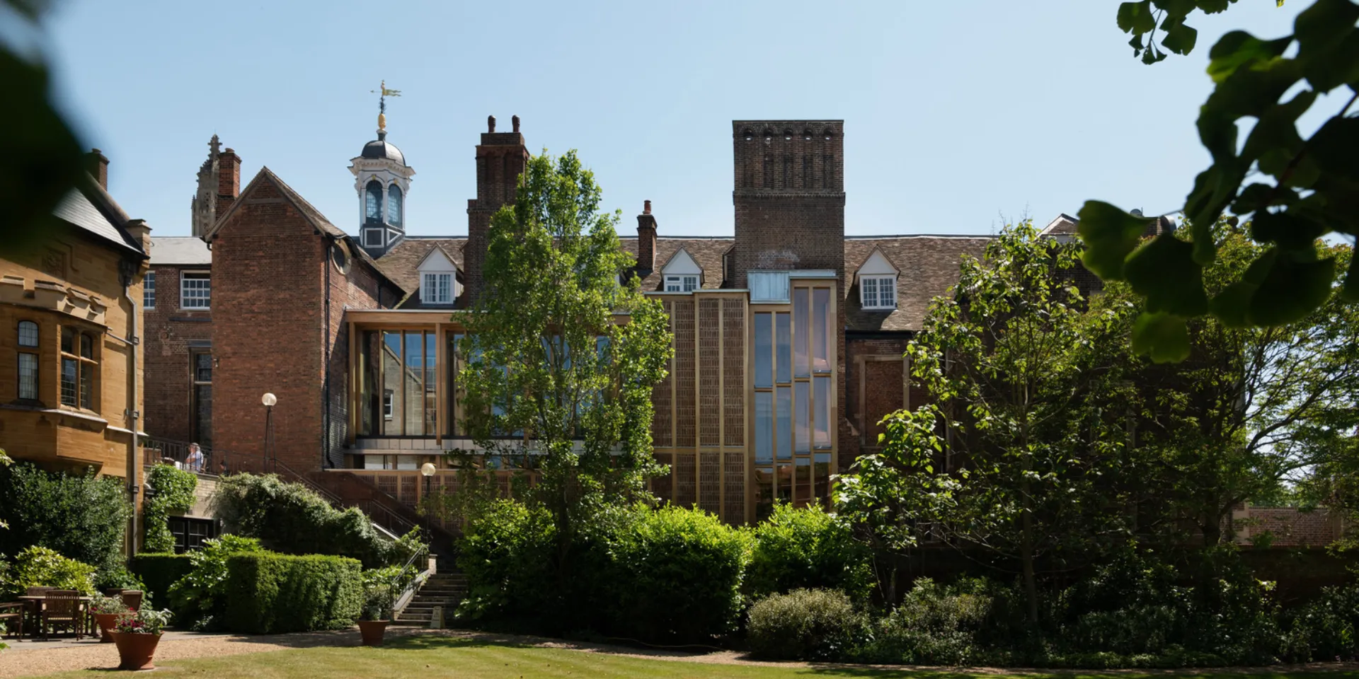The image shows a green lawn and trees with a brick building with a glass-panelled addition.