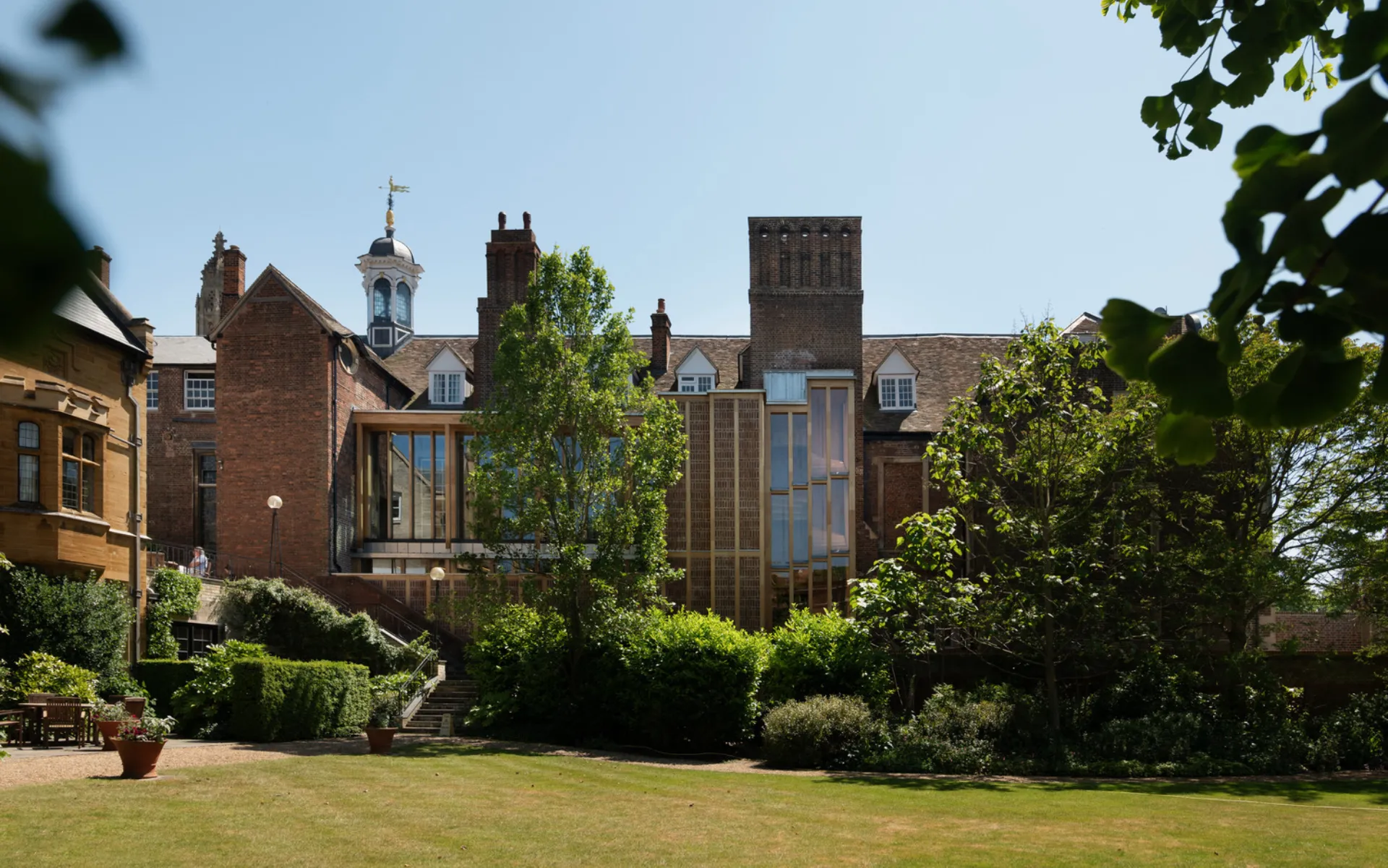 The image shows a green lawn and trees with a brick building with a glass-panelled addition.