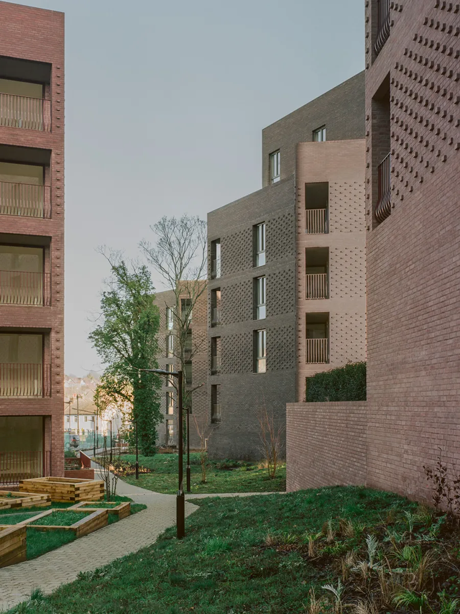 View through a landscaped courtyard between contemporary mid-rise residential buildings clad in textured red and dark brick. Balconies and recessed windows overlook winding paths, raised timber planters and newly planted greenery, with soft daylight highlighting the varied brickwork and garden spaces.