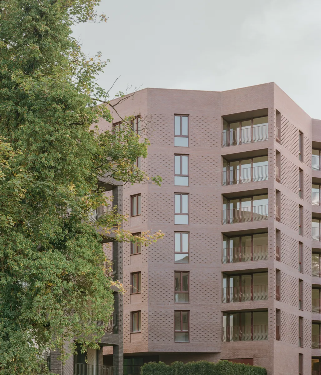 Mid-rise residential building clad in pale brick, featuring recessed balconies and textured brick detailing. Mature trees frame the building in the foreground, with greenery contrasting against the geometric façade under an overcast sky.