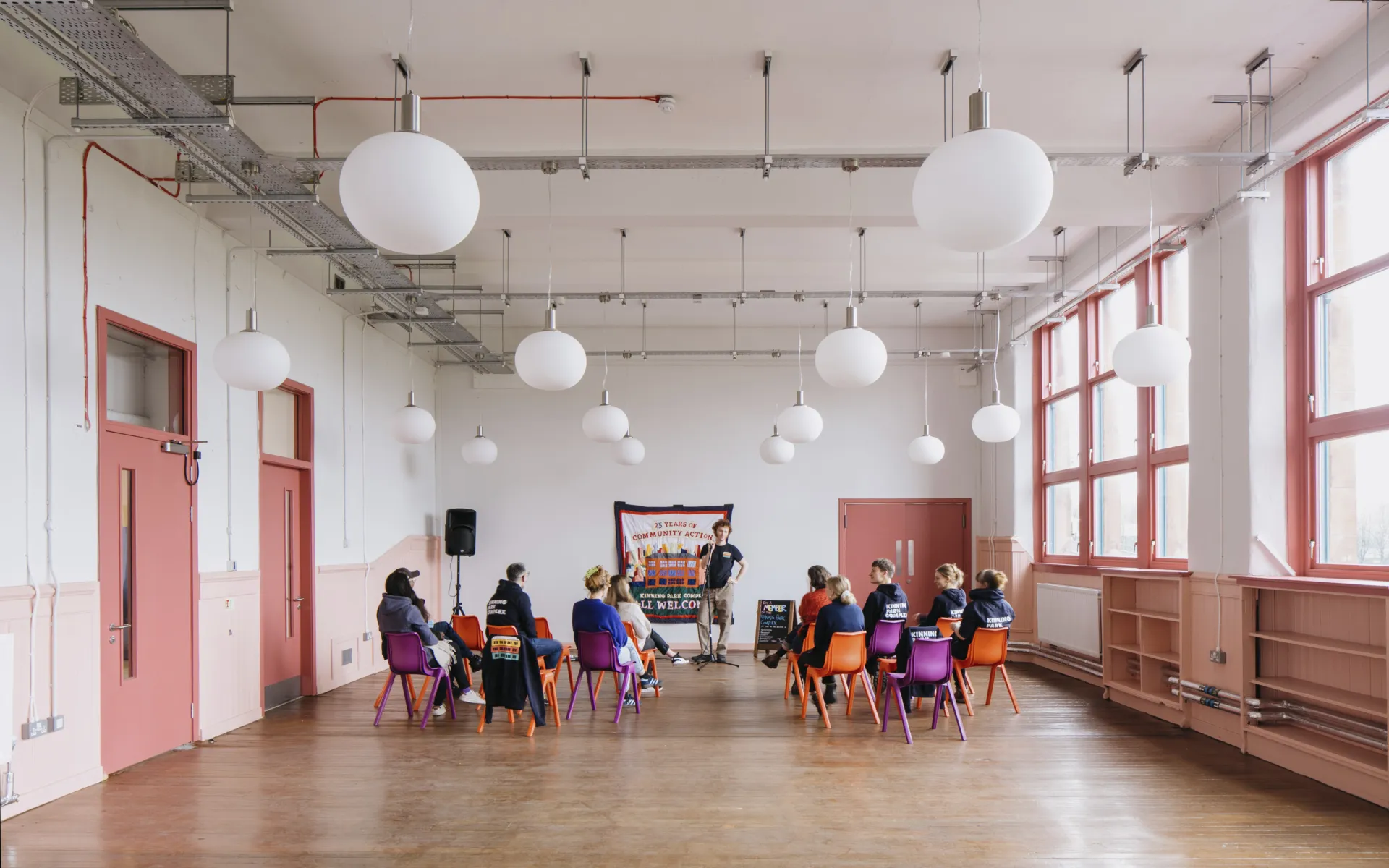 A large wooden-floored room lit with daylight from large windows, with several people sitting in small groups