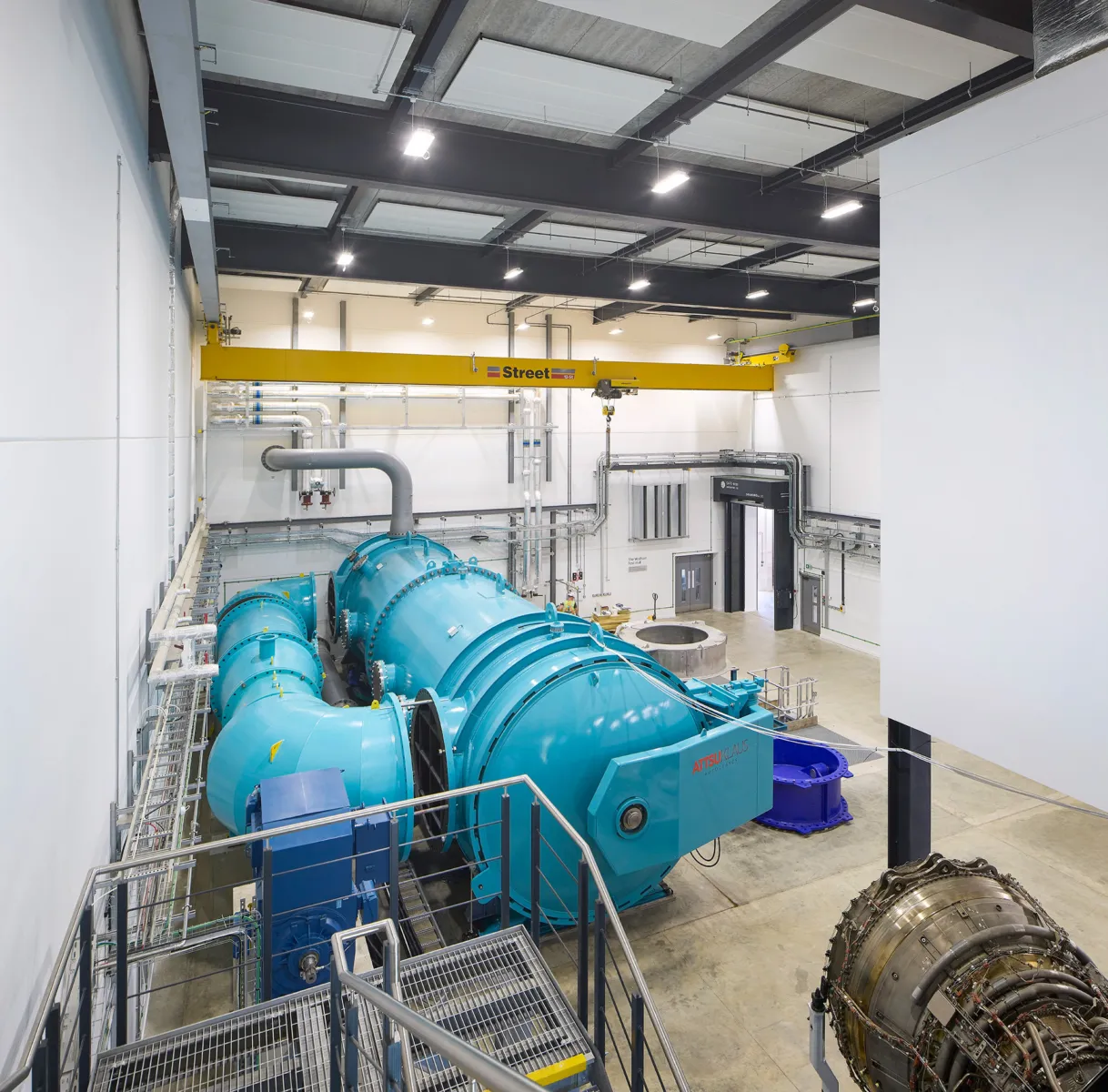 Large laboratory testing hall containing a blue industrial research rig connected to extensive pipework and services. An overhead gantry crane spans the space, with access platforms and specialist equipment visible throughout.