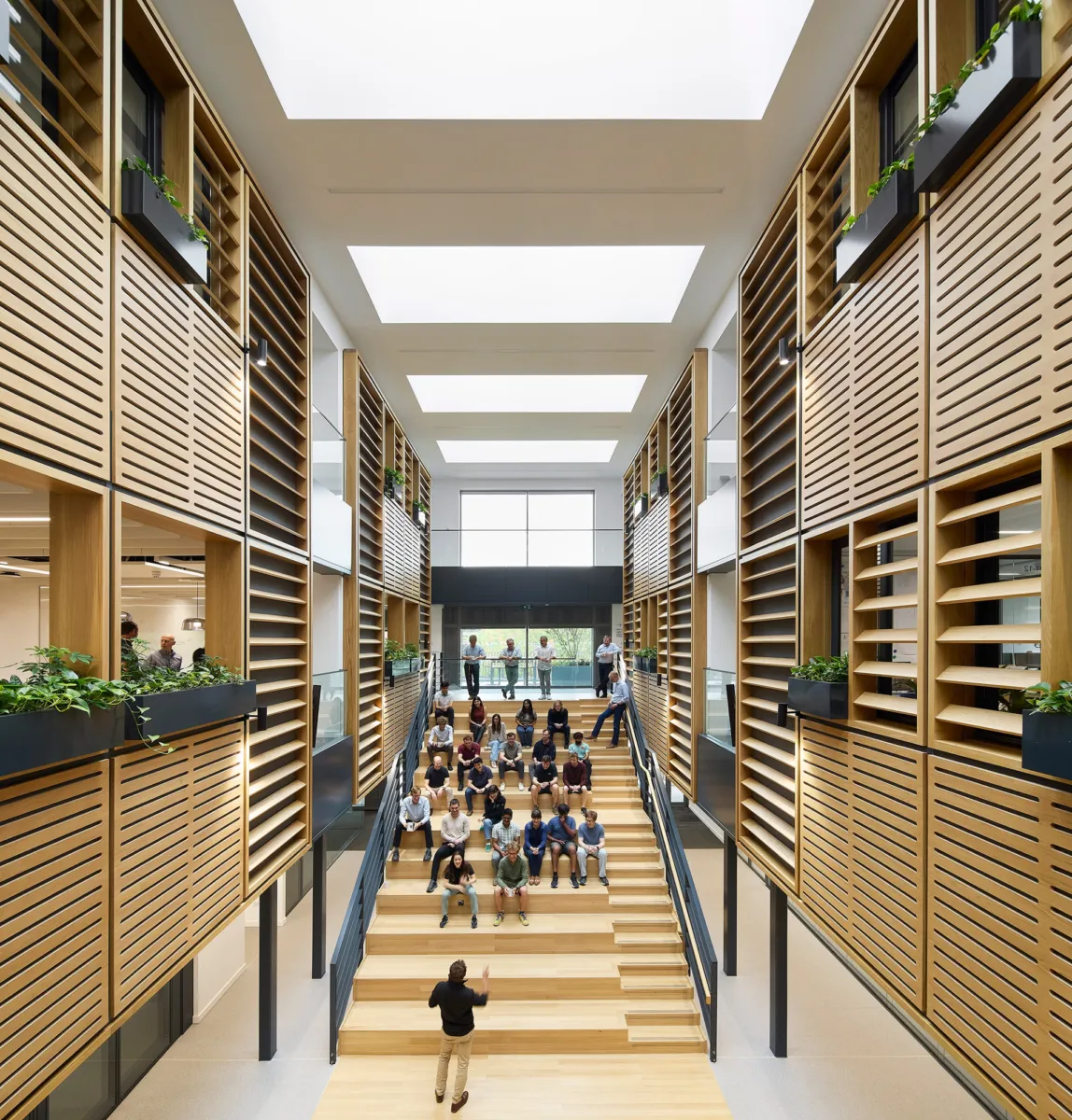 Multi-storey atrium featuring a wide timber social staircase used as informal seating, framed by timber louvred walls, integrated planters and large rooflights. A group of people are gathered on the steps while others stand on the surrounding balconies.