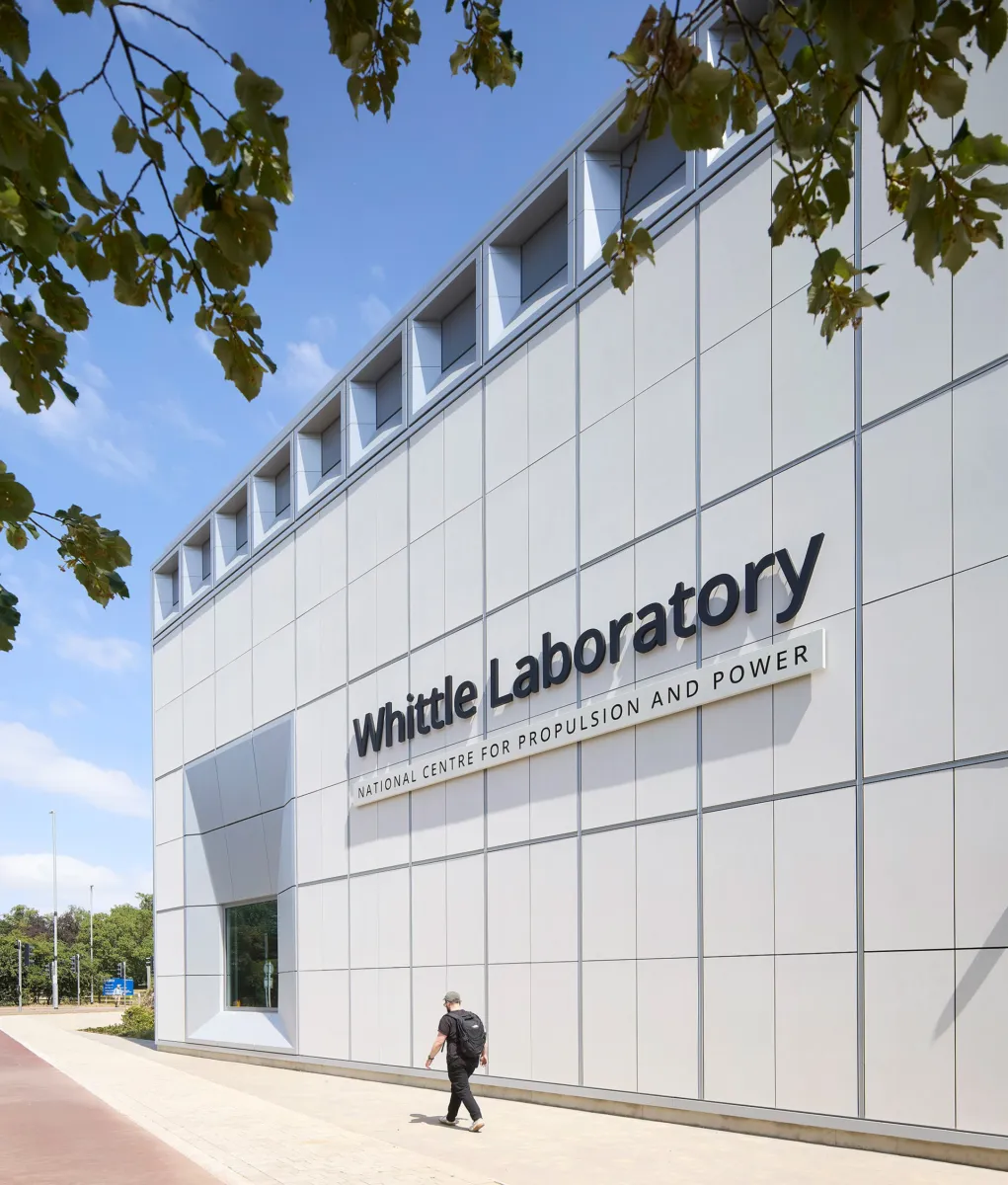 Side elevation of the Whittle Laboratory building, featuring a white metal-clad façade with recessed windows and large signage reading “Whittle Laboratory – National Centre for Propulsion and Power”. A pedestrian walks along the pavement beneath tree branches framing the view.