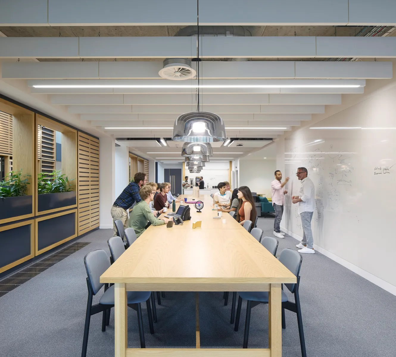 Open-plan collaboration area with people gathered around a long communal table, while others use a whiteboard wall for discussion. The space features timber detailing, integrated planting, suspended lighting and exposed services.