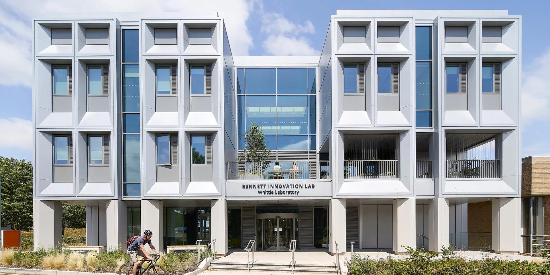 Front elevation of the Bennett Innovation Lab at the Whittle Laboratory in Cambridge, showing a symmetrical white façade with deep-set windows, a glazed central entrance and landscaped planting. A cyclist passes in the foreground.