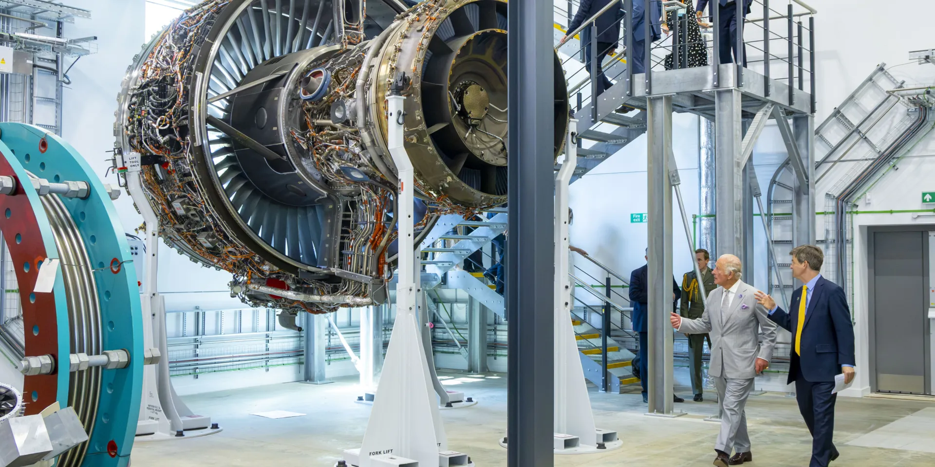 Large aircraft engine displayed inside a research and testing facility, showing exposed turbine blades, wiring and mechanical components. Several visitors, including King Charles III, are walking around the laboratory and viewing the engine from an elevated steel access platform, while additional testing equipment and industrial services are visible throughout the bright, high-ceilinged space.