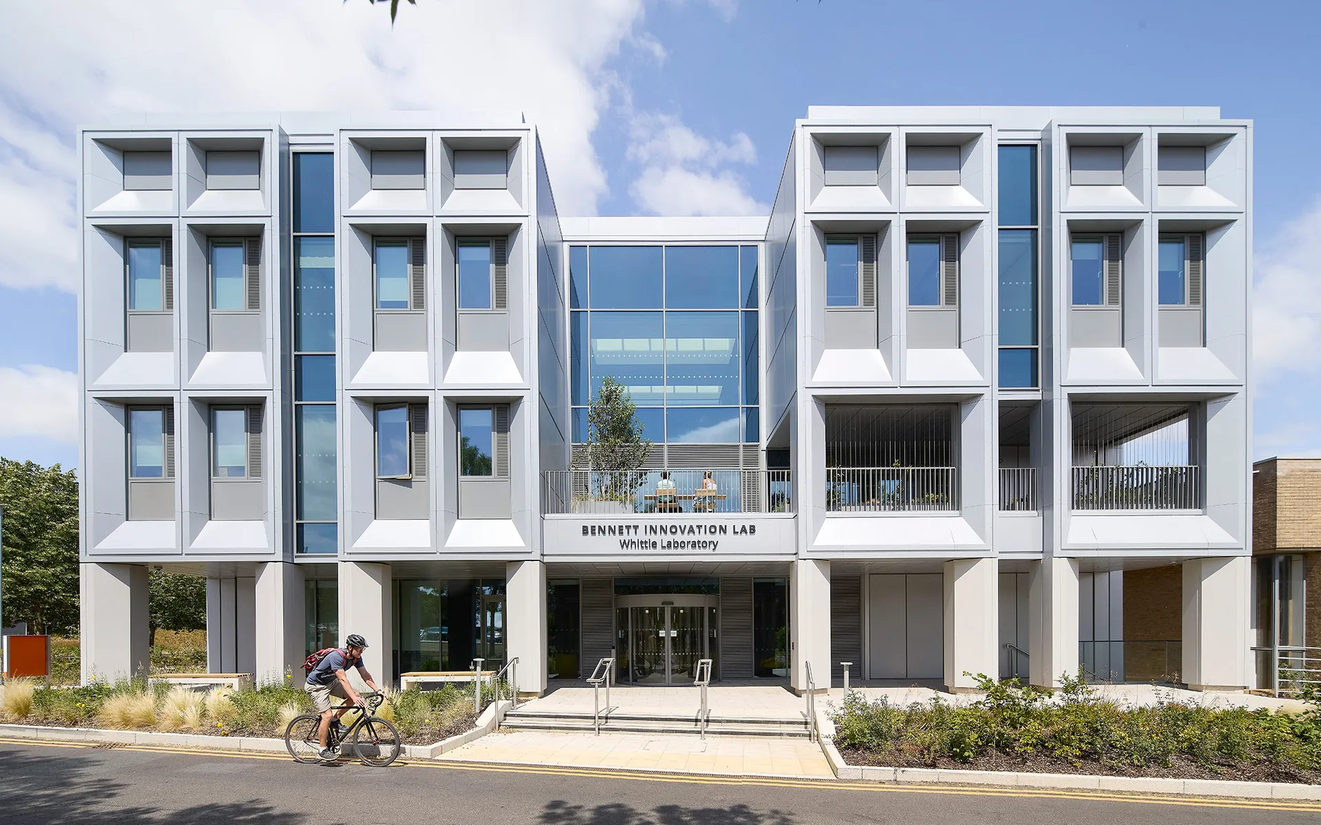 Front elevation of the Bennett Innovation Lab at the Whittle Laboratory in Cambridge, showing a symmetrical white façade with deep-set windows, a glazed central entrance and landscaped planting. A cyclist passes in the foreground.