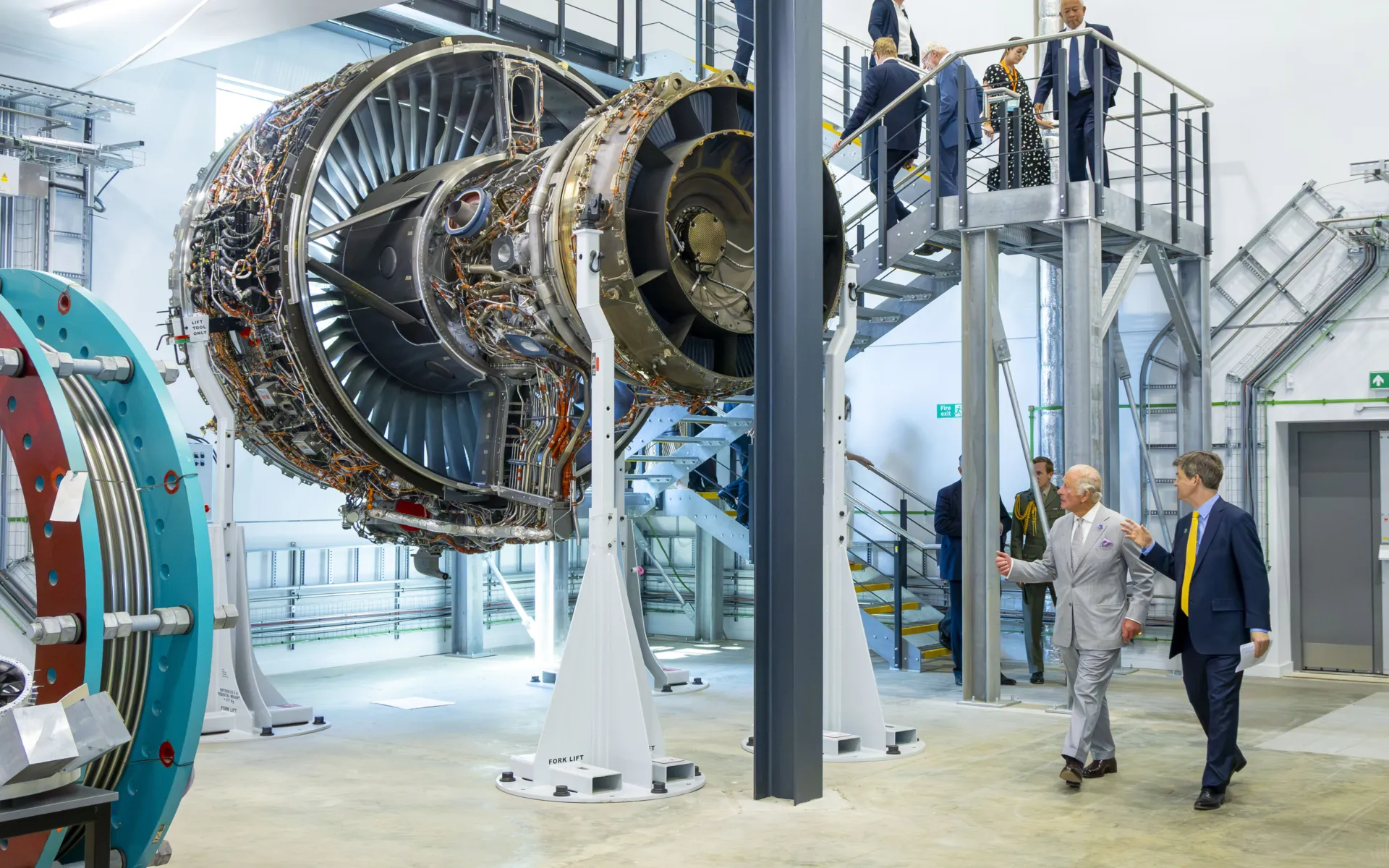 Large aircraft engine displayed inside a research and testing facility, showing exposed turbine blades, wiring and mechanical components. Several visitors, including King Charles III, are walking around the laboratory and viewing the engine from an elevated steel access platform, while additional testing equipment and industrial services are visible throughout the bright, high-ceilinged space.
