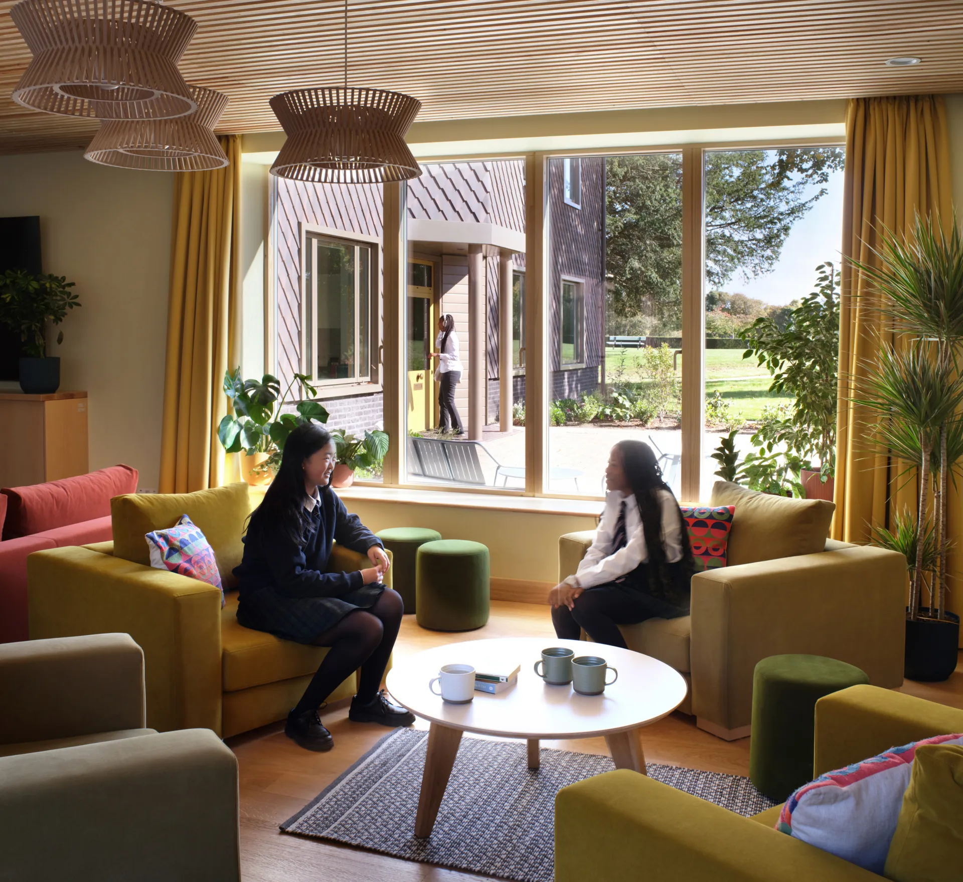 Two girls sat chatting in armchairs, with a coffee table between them, in a living room. A window behind them looks out across a courtyard.