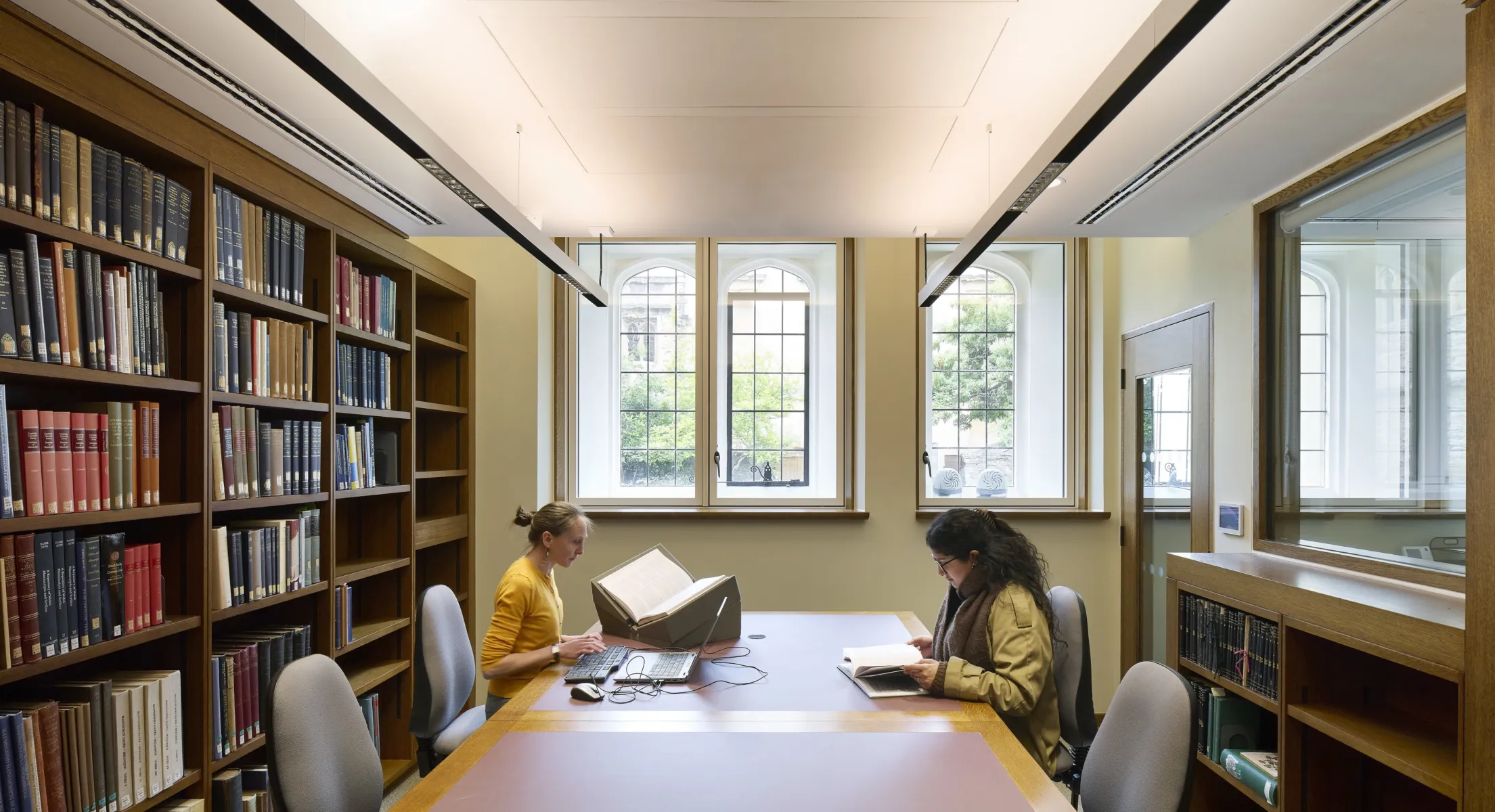 Two people are studying in a modern-looking library. Books line the shelves and there is a modern window with a historic window behind it.