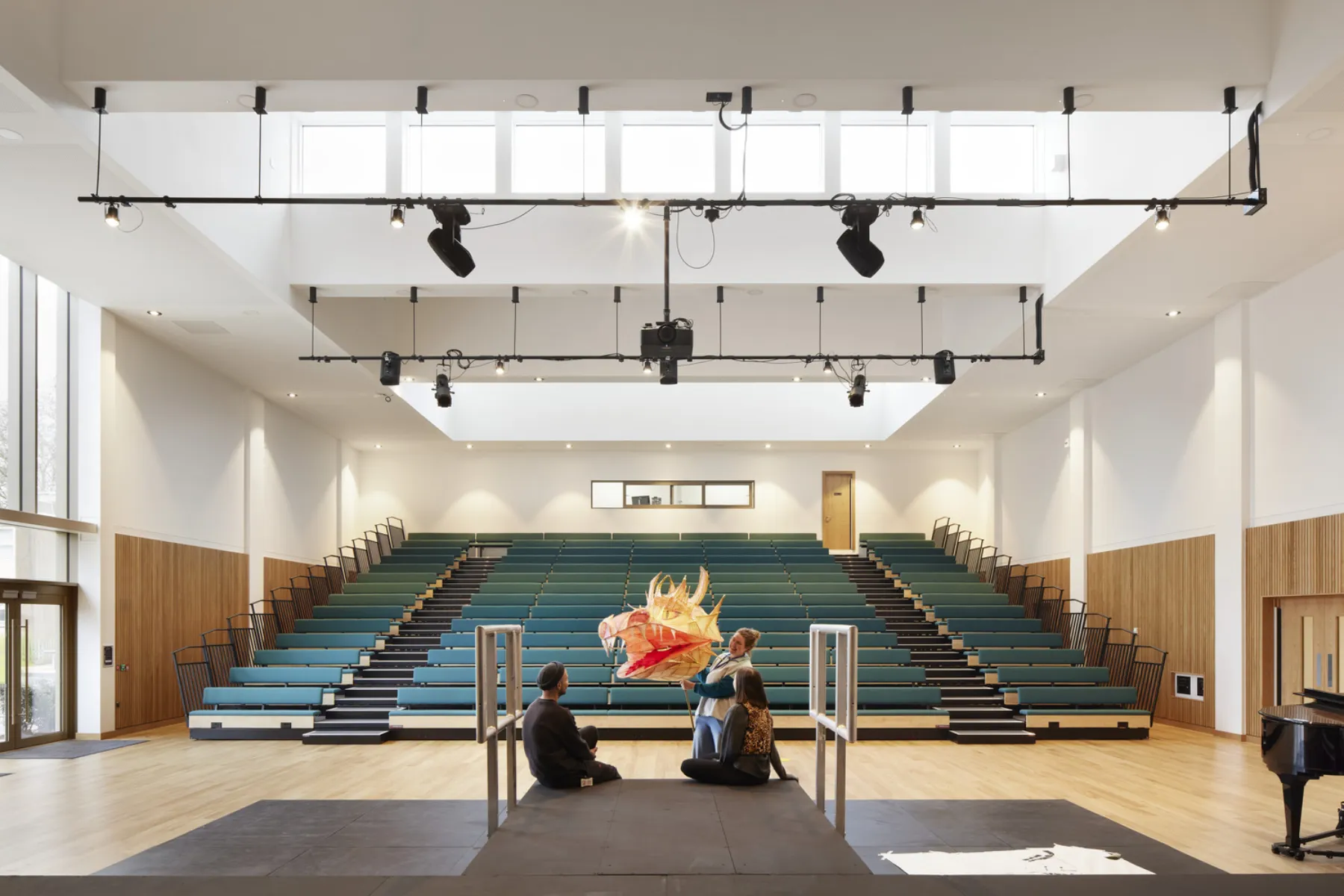 Large performance hall with tiered seating, overhead lighting rig and three people gathered on a stage with a dragon puppet.
