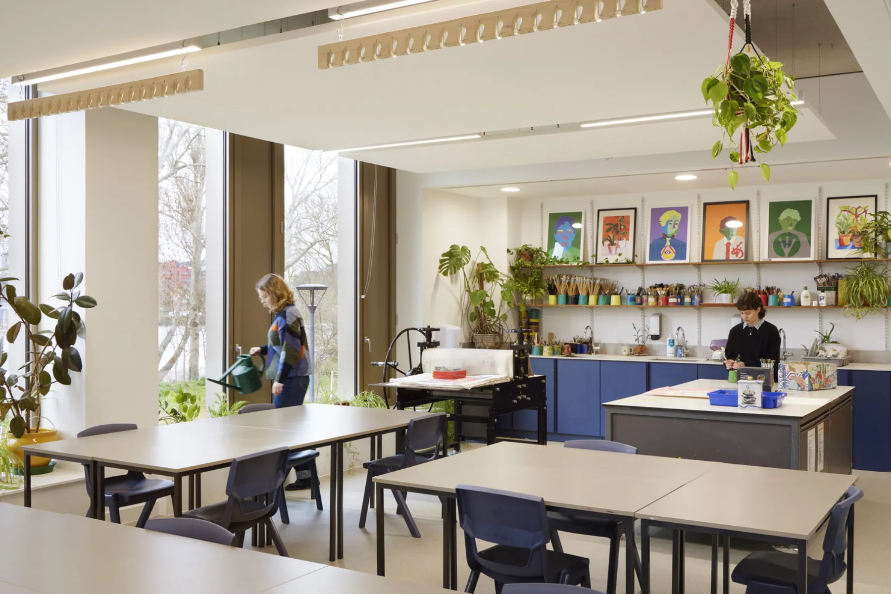 Bright art classroom with worktables, plants, printmaking equipment and students working near large windows.