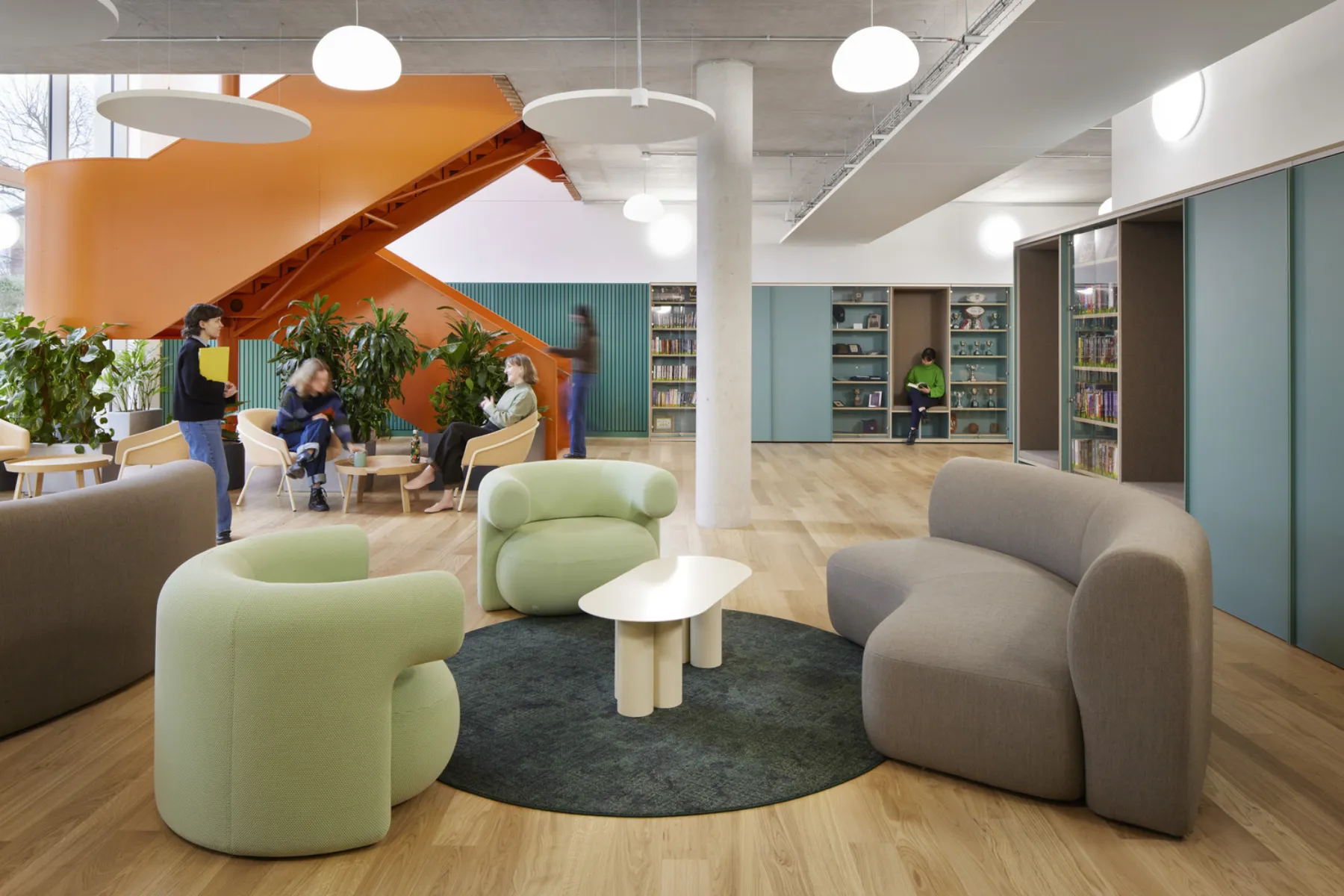 Library lounge with soft seating, bookshelves and an orange feature staircase, with people reading and meeting.