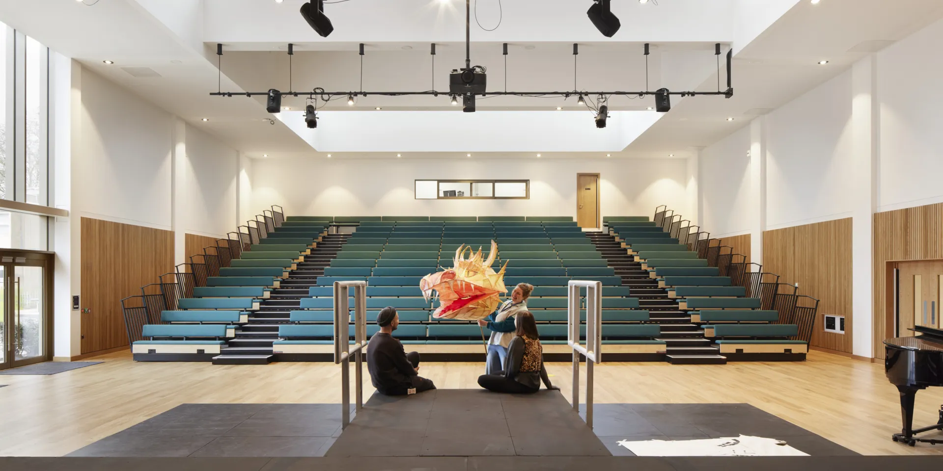 Large performance hall with tiered seating, overhead lighting rig and three people gathered on a stage with a dragon puppet.
