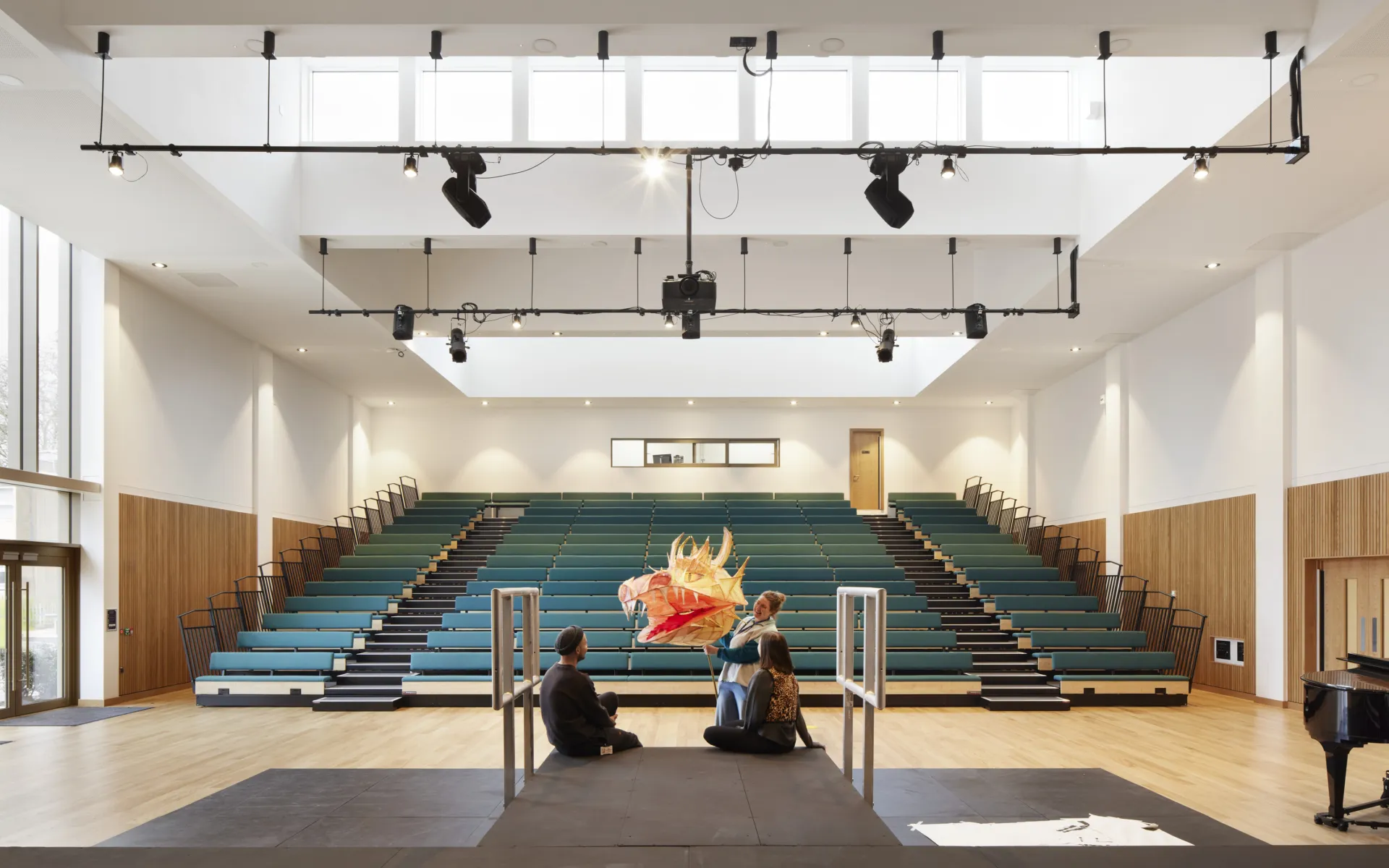 Large performance hall with tiered seating, overhead lighting rig and three people gathered on a stage with a dragon puppet.