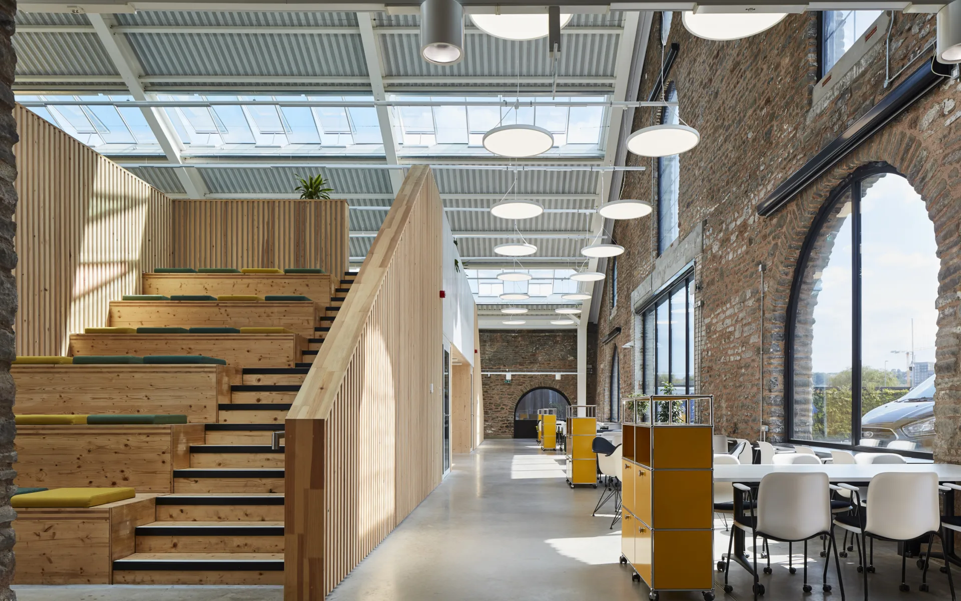image shows a large wooden hellerup stair case which allows for seating as well as stairs. The image shows a brick wall with large windows as well as and a ceiling with skylights.