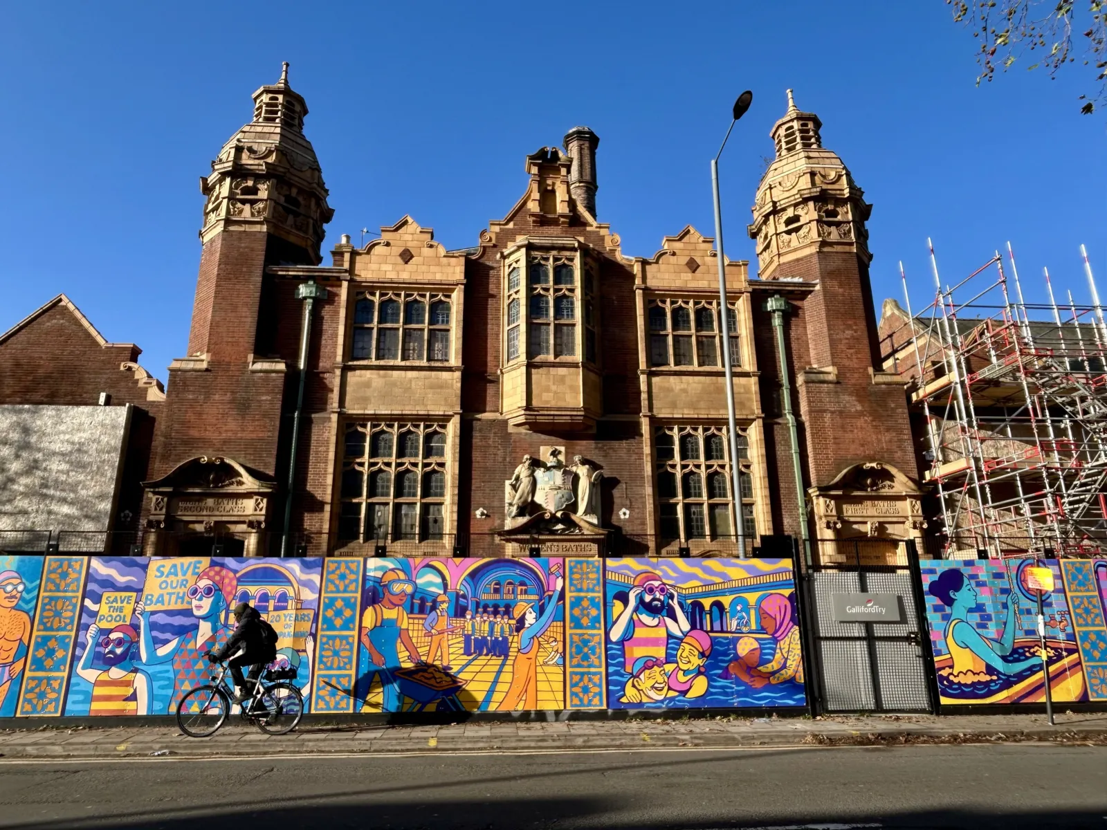 Red brick historic bathhouse with ornate towers, partially hidden behind colourful illustrated hoarding; cyclist passing in front.