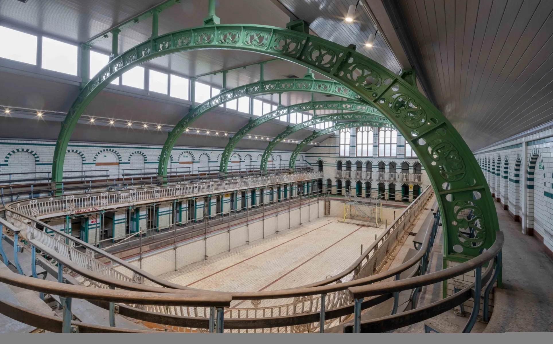 Empty historic gala pool hall with tiled basin, balconies, and large green iron roof arches.