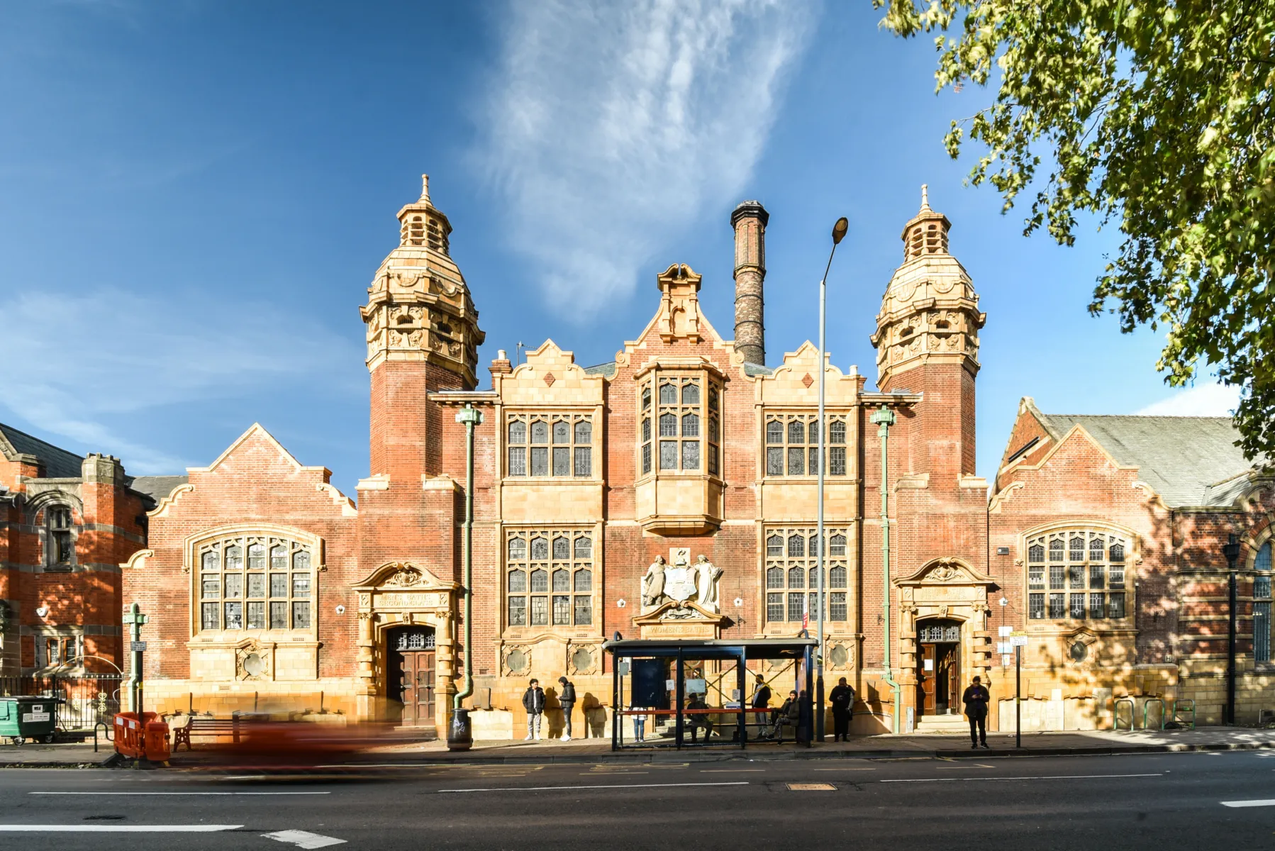 Symmetrical red brick bathhouse with twin towers, seen across the street with bus stop and people in front.