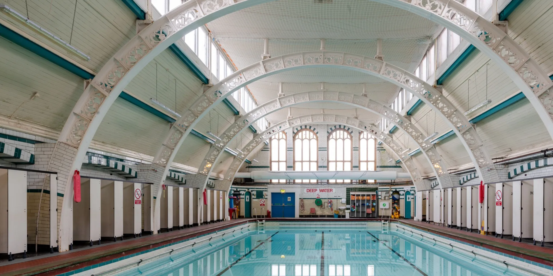 Restored indoor pool with still water, tiled walls, and decorative white arch trusses overhead.