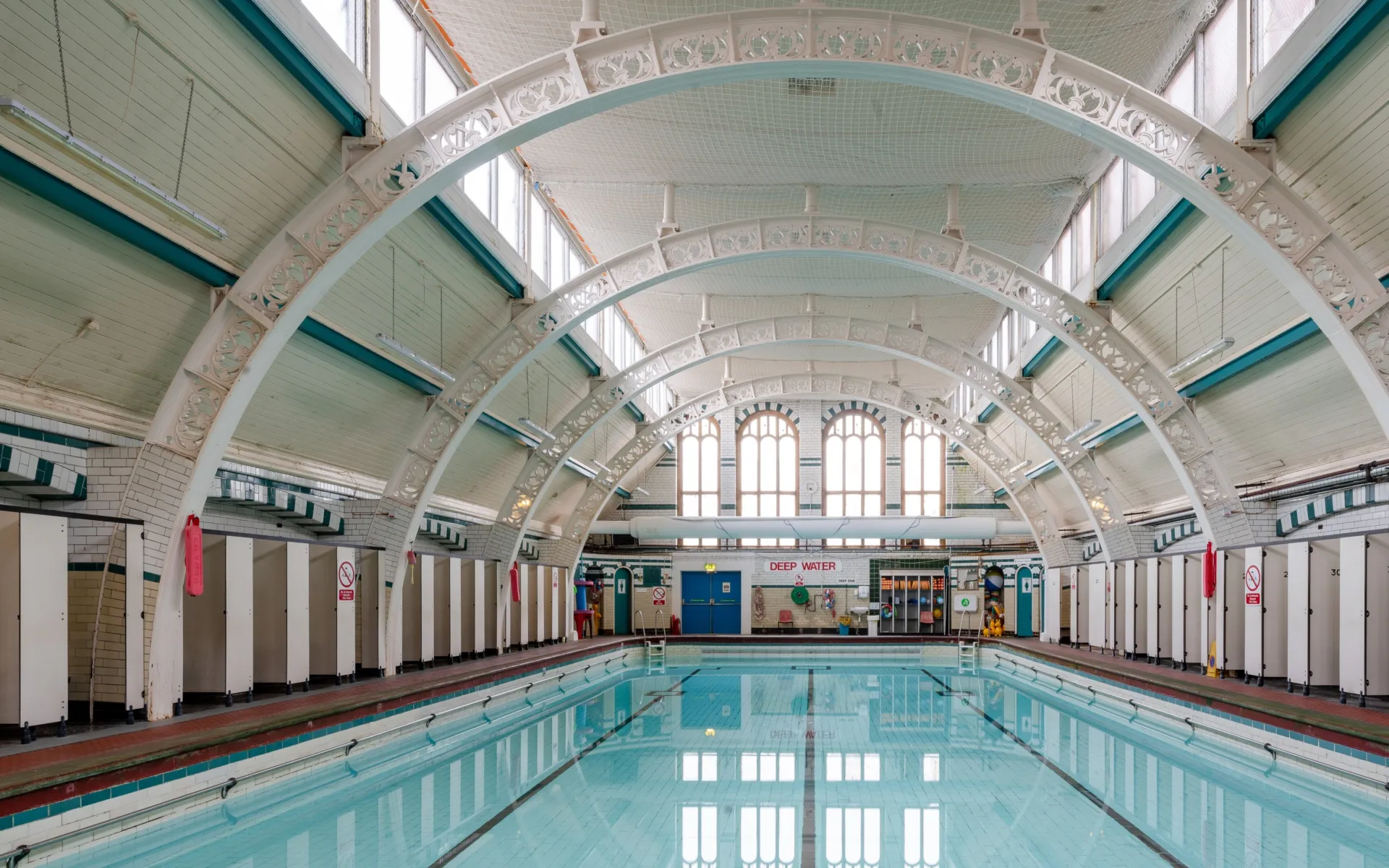 Restored indoor pool with still water, tiled walls, and decorative white arch trusses overhead.