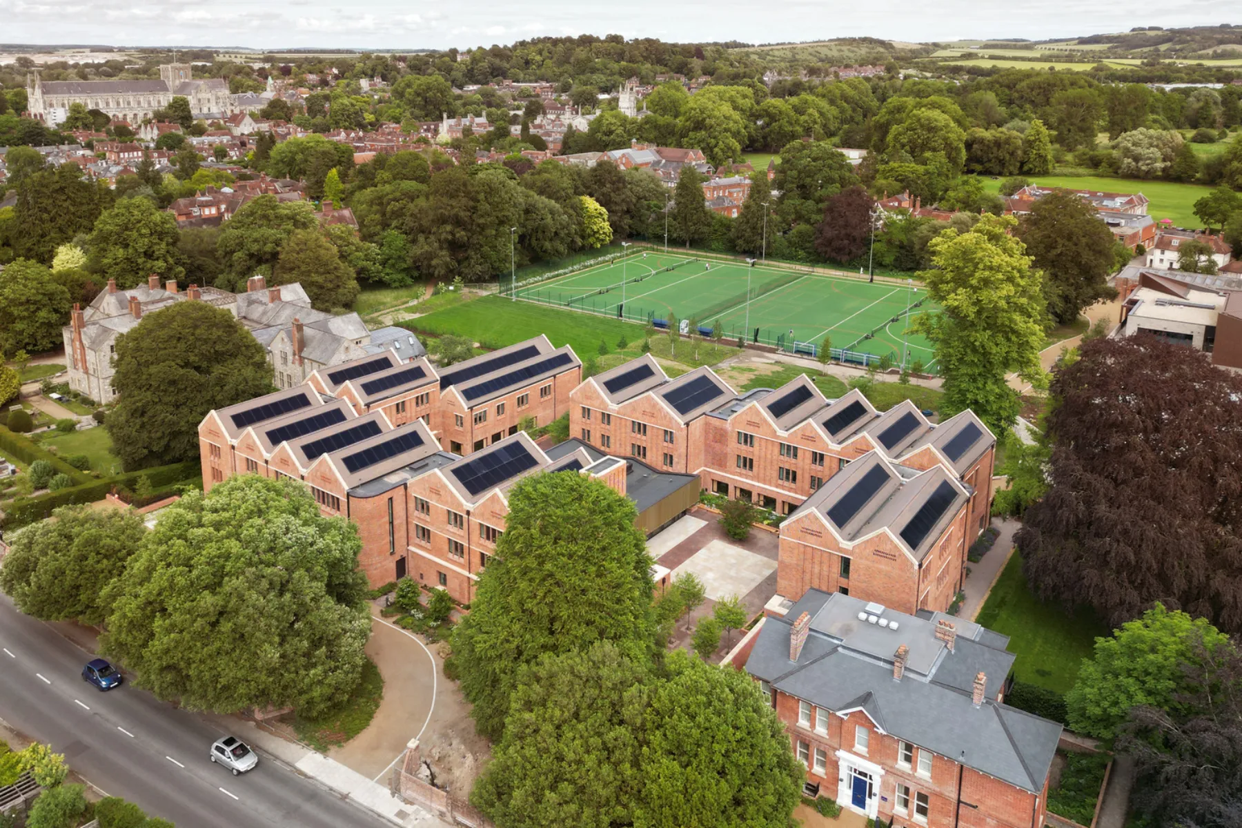 Aerial view of Winchester College’s new brick boarding houses arranged around courtyards, with rooftop solar panels, sports pitches and the surrounding landscape visible beyond.