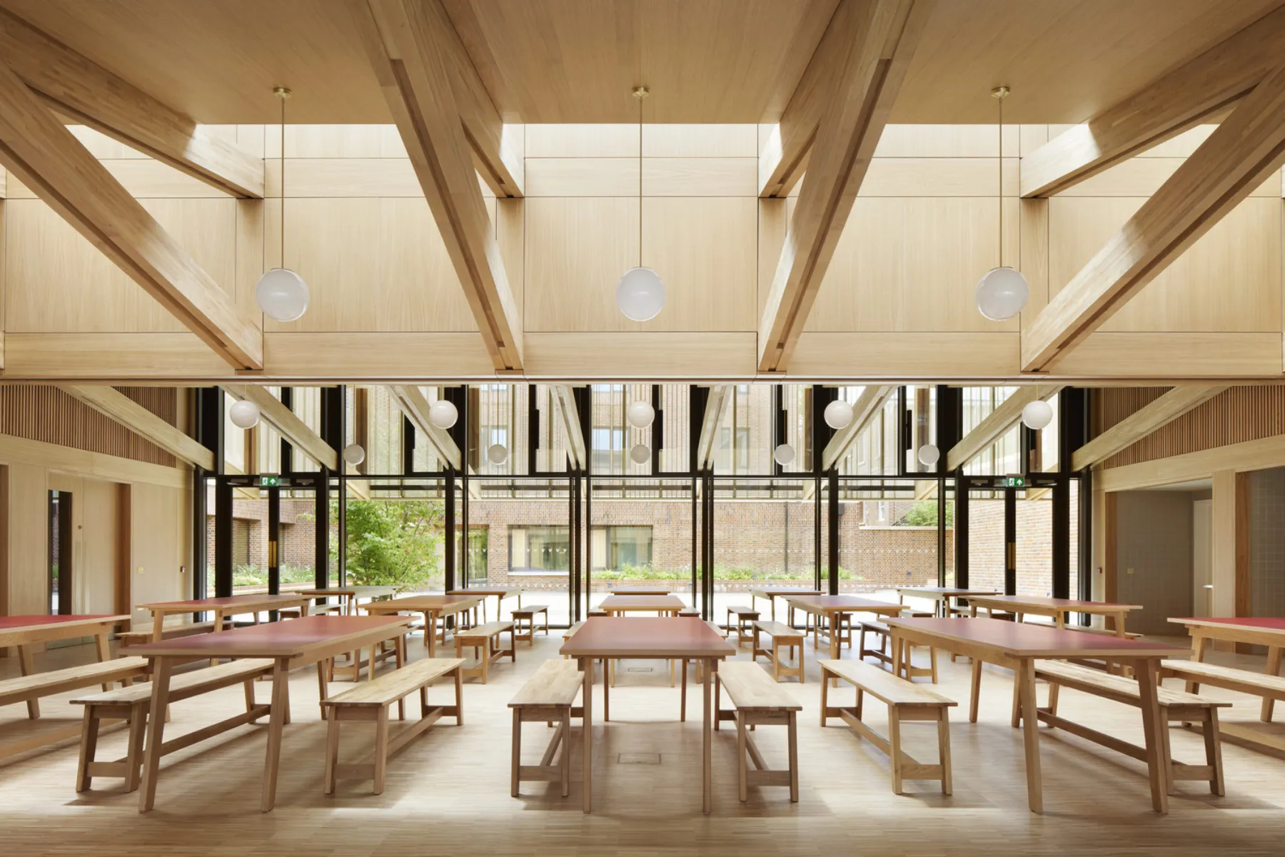Timber-lined dining hall with long communal tables and benches beneath exposed timber beams and pendant lighting, opening onto a landscaped courtyard.