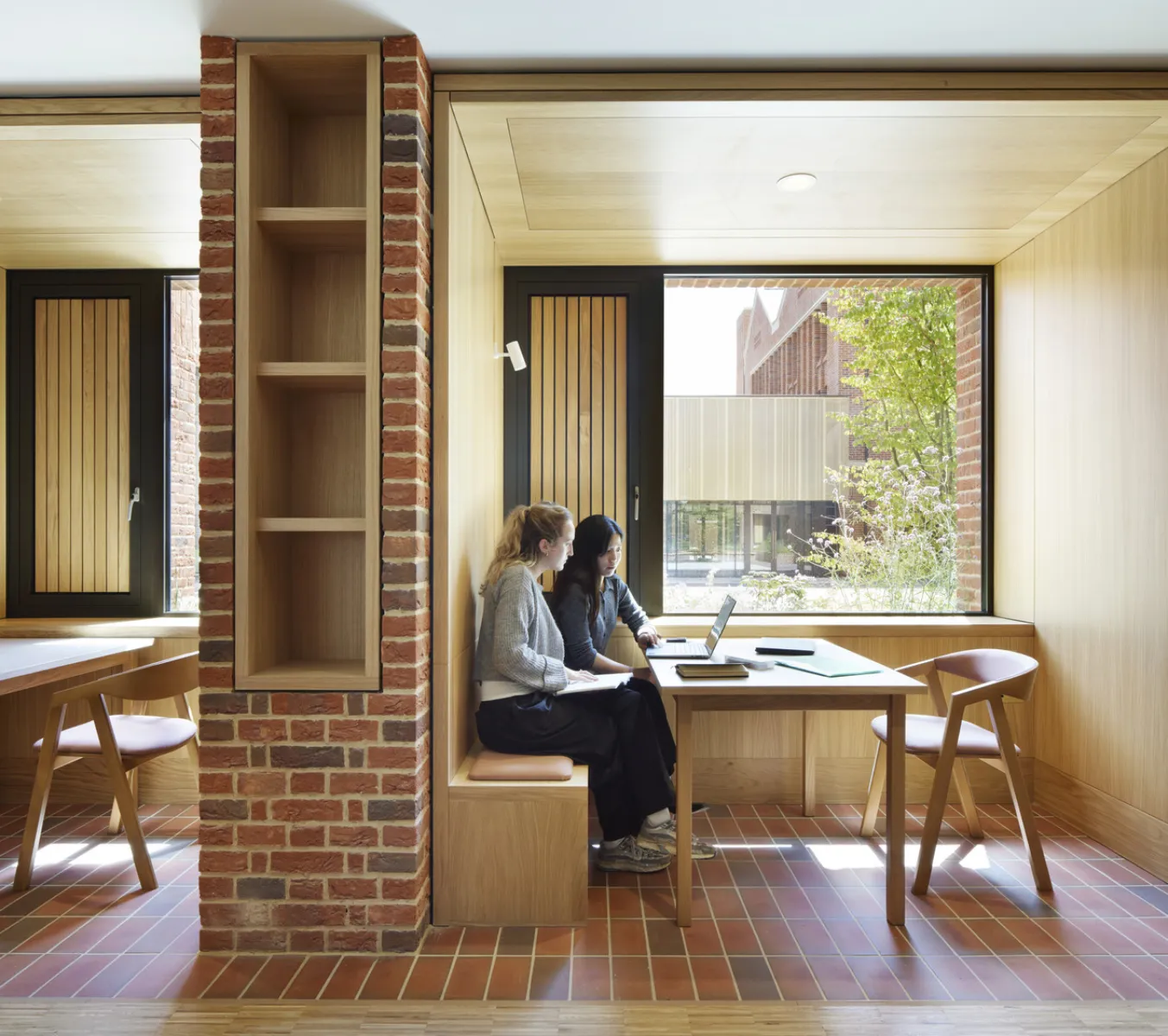 Two students seated at a built-in study booth within a timber-lined interior, working at a table beside a large window overlooking the courtyard.