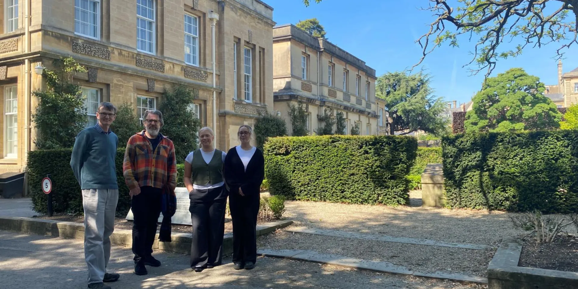 Two men and two women stand in front of a sandstone historic building at Magdalen College, Oxford