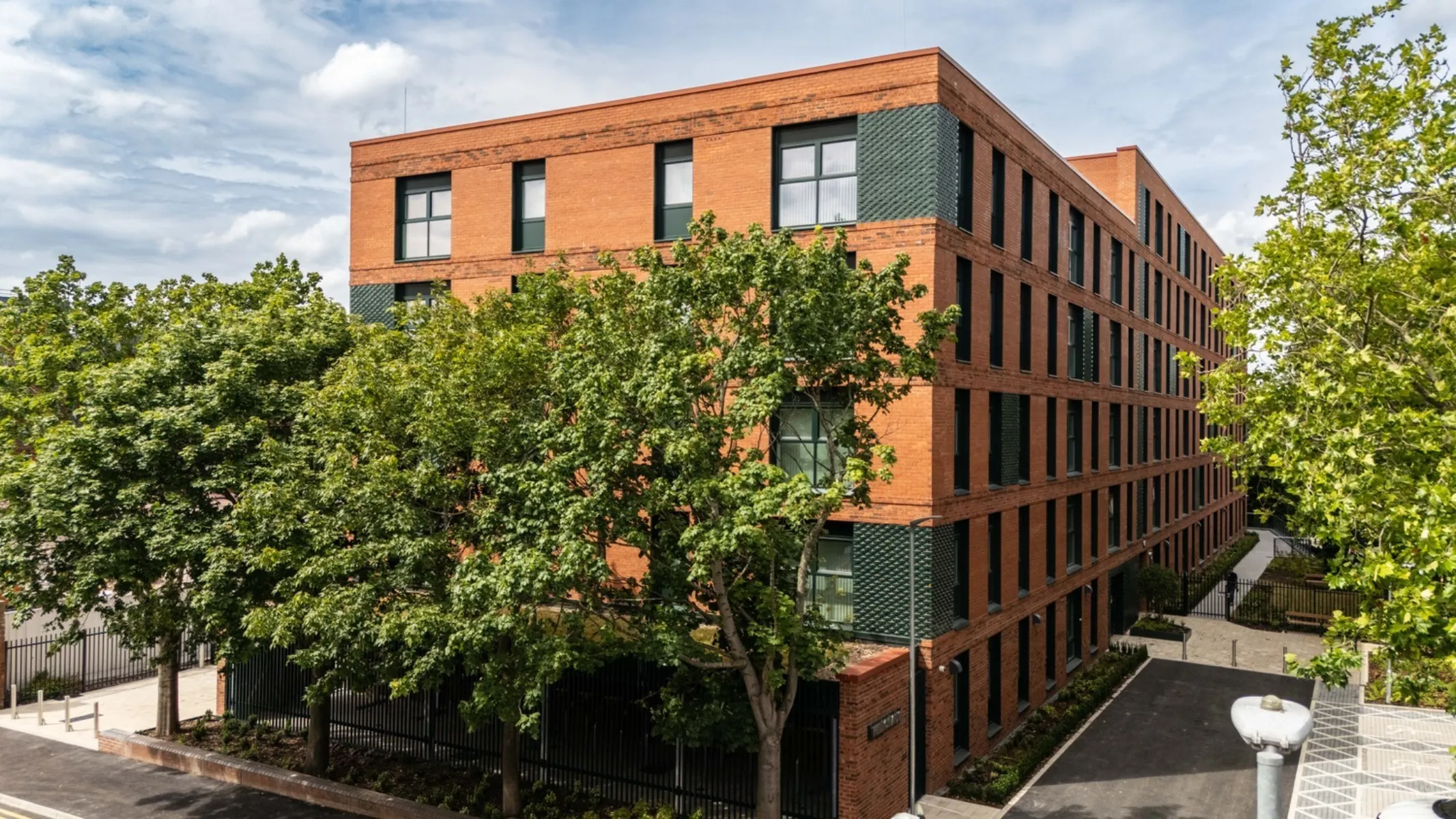 Contemporary red brick residential building with a regular pattern of tall windows and dark textured brick detailing. Mature trees partially screen the façade, with landscaped pathways and a courtyard surrounding the building under a bright, cloudy sky.